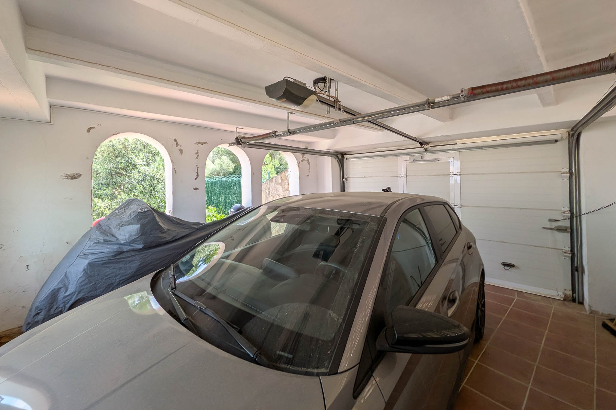 Afgesloten garage voorzien van een automatische sectionaaldeur, tegelvloer en balkenplafond. Boogopeningen zorgen voor natuurlijke lichtinval en ventilatie.