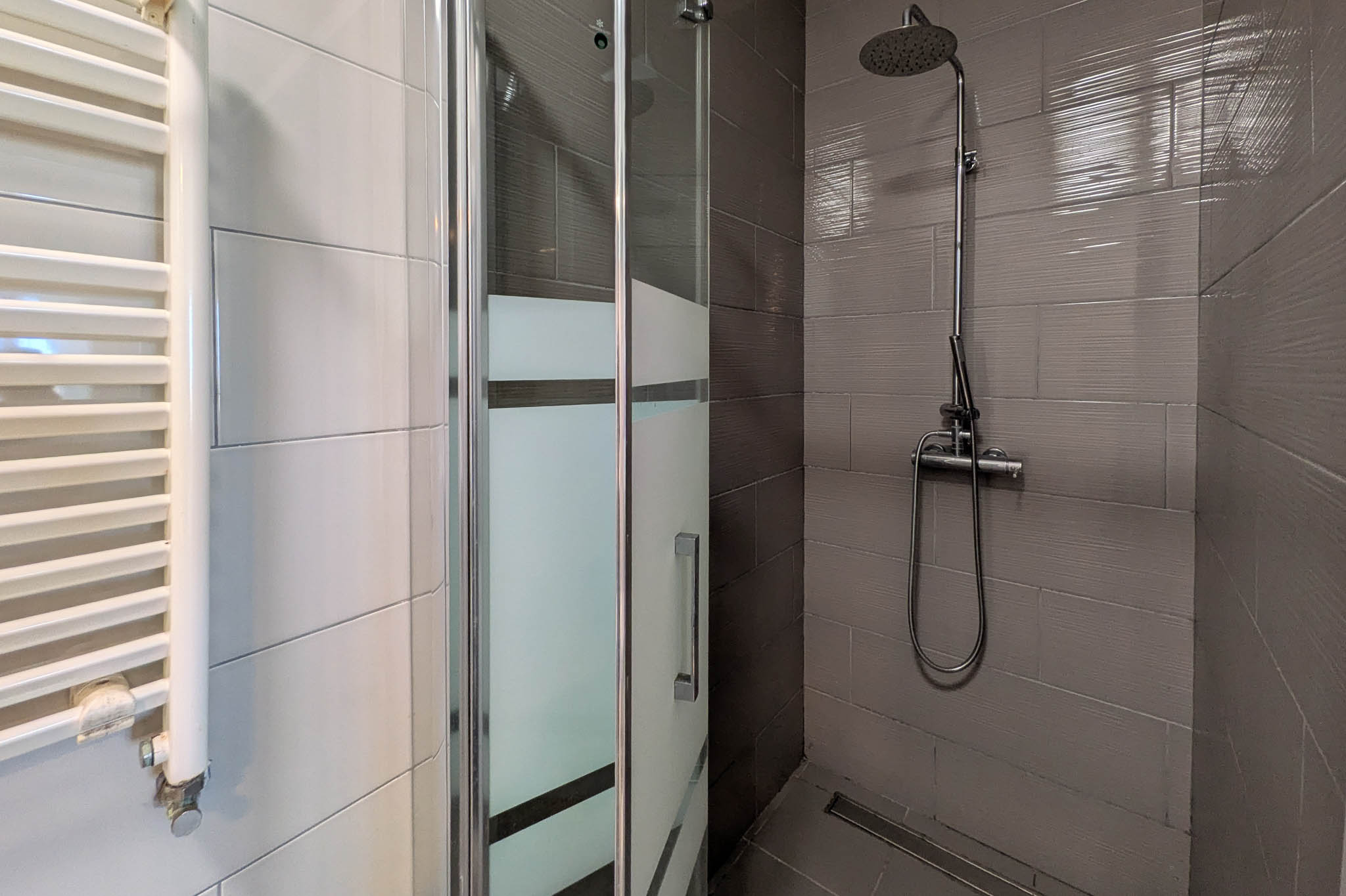 Badkamer met inloopdouche voorzien van grijze structuurtegels, glazen wand met matte banen, chromen doucheset en een witte handdoekradiator.