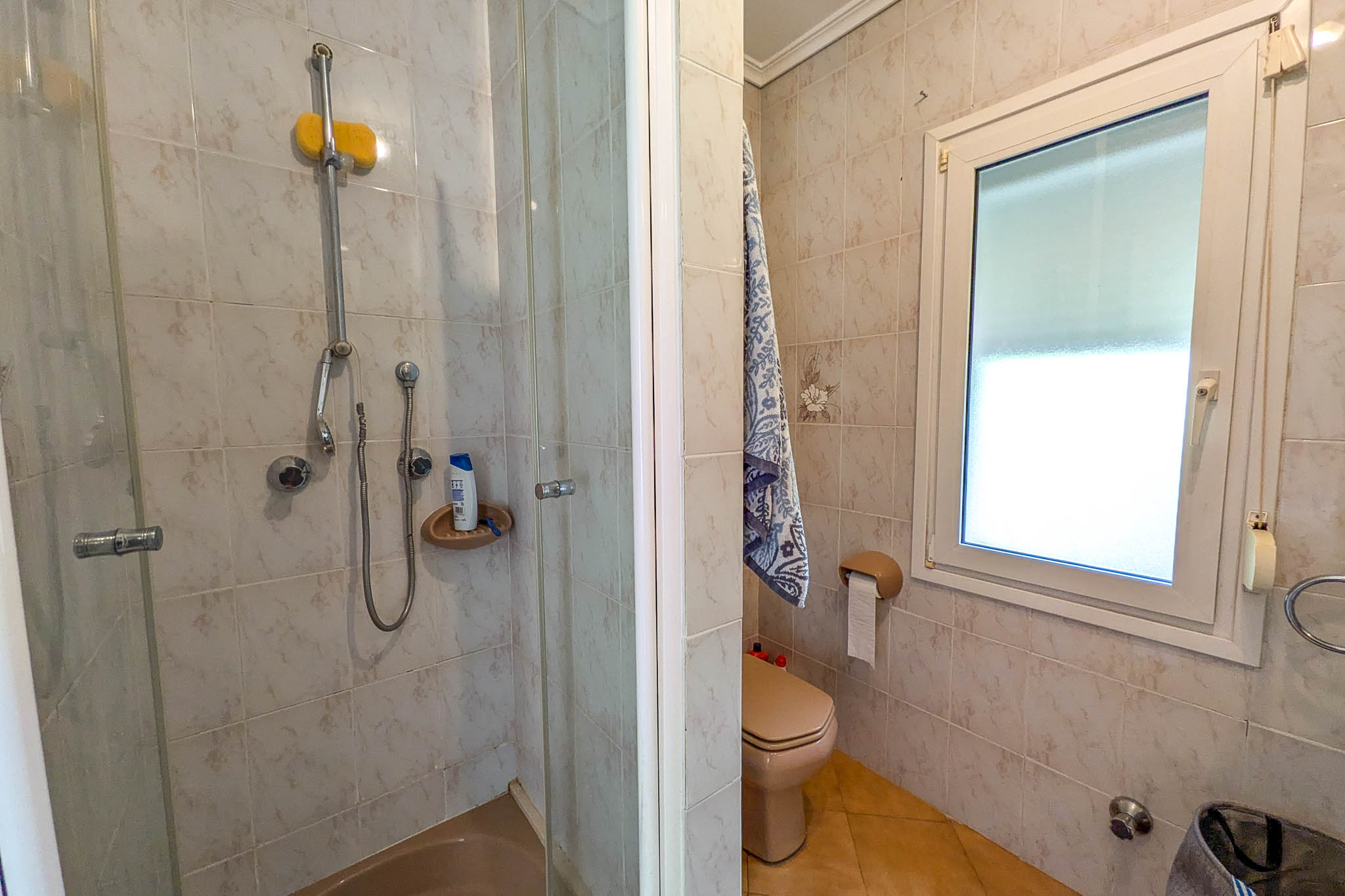 Badkamer voorzien van een glazen douchecabine, crèmekleurig toilet en een wit PVC-raam voor daglicht. De wanden zijn afgewerkt met gedecoreerde keramische tegels.
