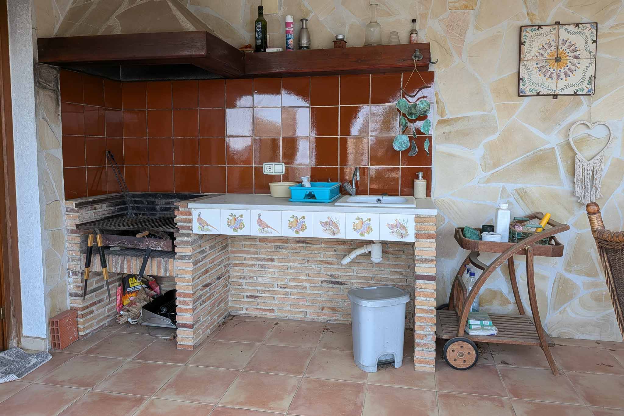 Buitenkeuken met ingebouwde stenen barbecue, spoelbak en decoratief betegeld werkblad tegen een wand van natuursteen.