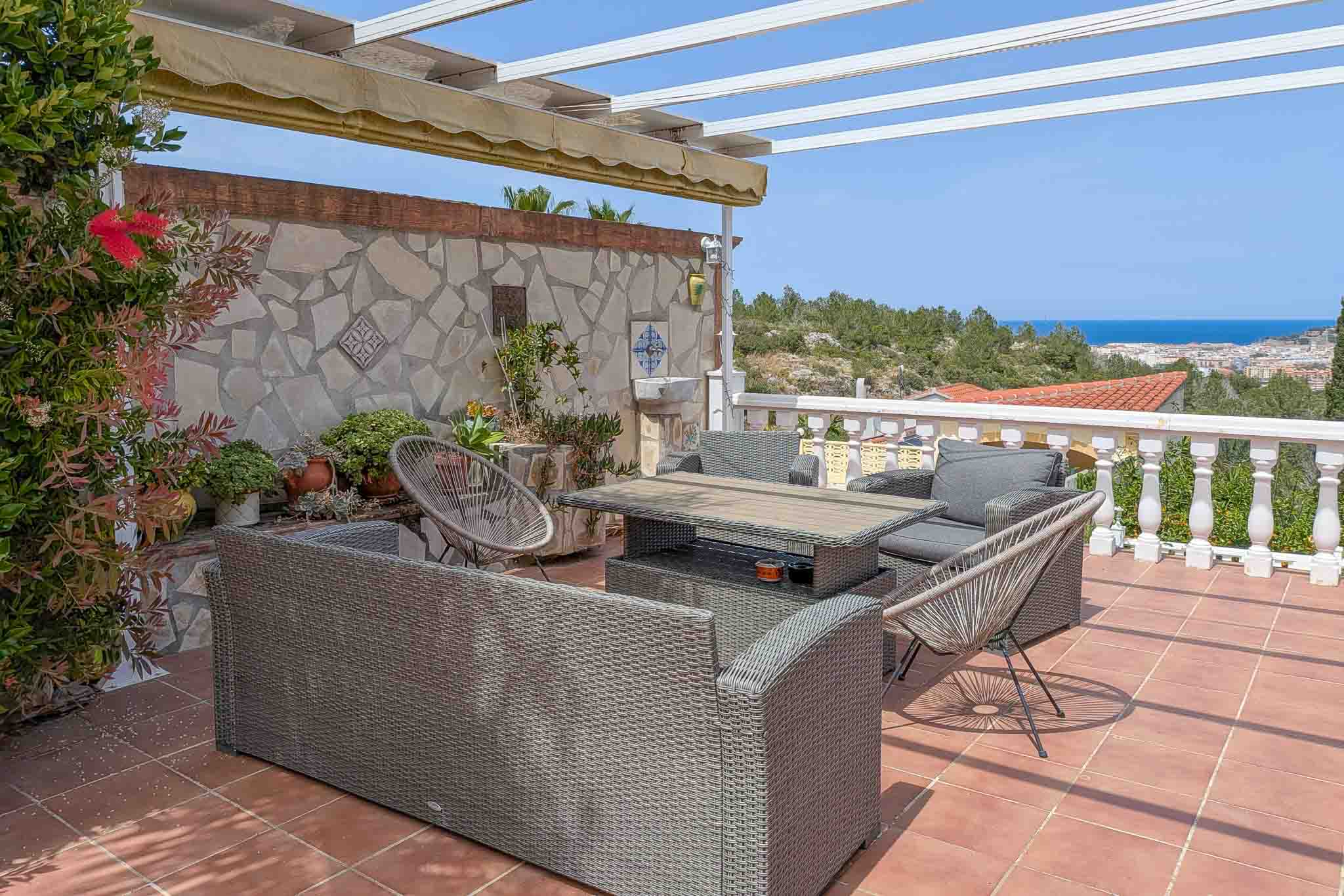 Buitenterras met rotan meubilair, terracotta vloer en panoramisch uitzicht op zee. Voorzien van een pergola met zonnescherm en een decoratieve natuurstenen muur.