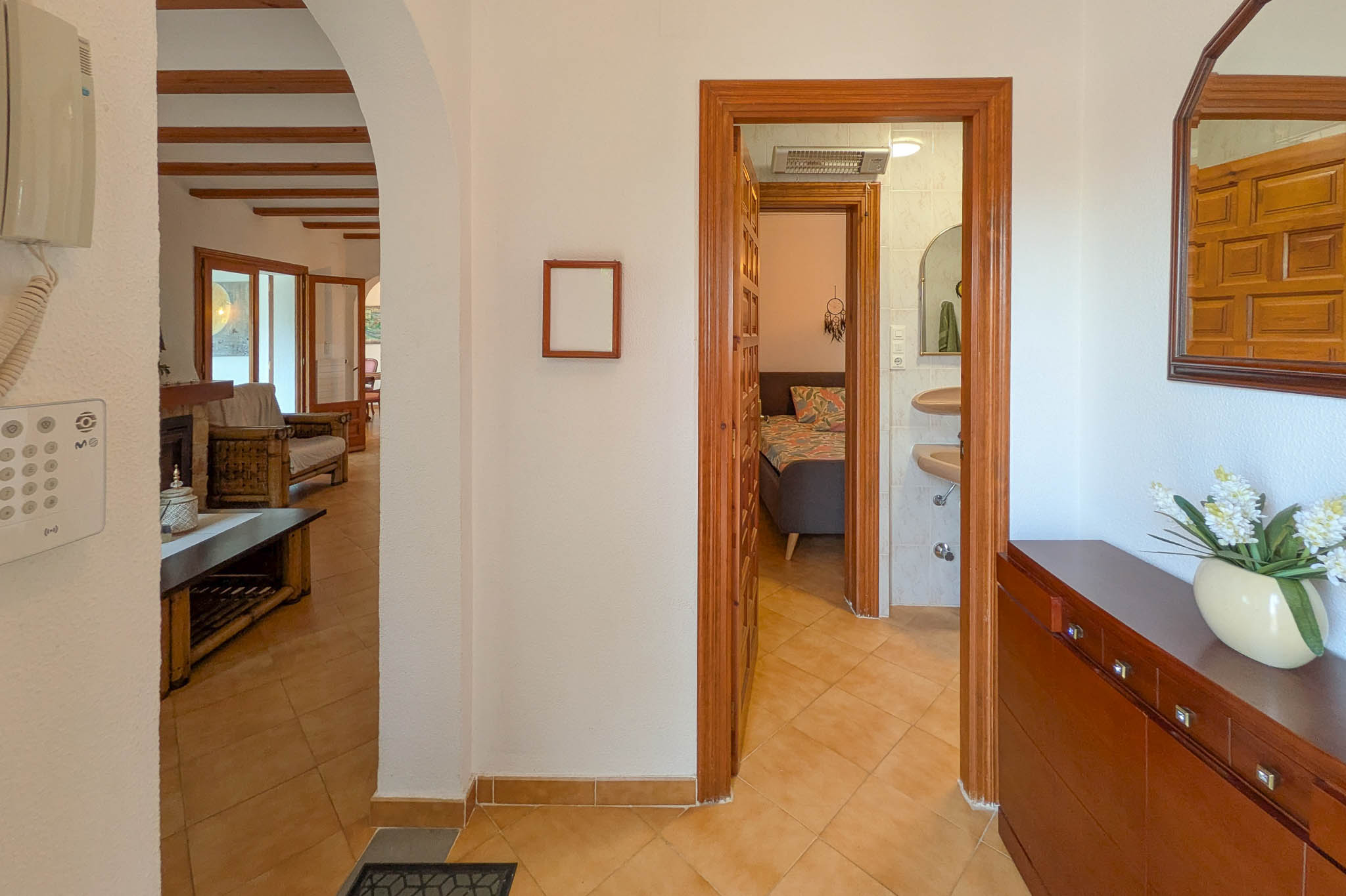 Hal met terracotta vloertegels die de woonkamer met houten balken verbindt met een slaapkamer en badkamer, afgewerkt met houten kozijnen en een klassieke boog.