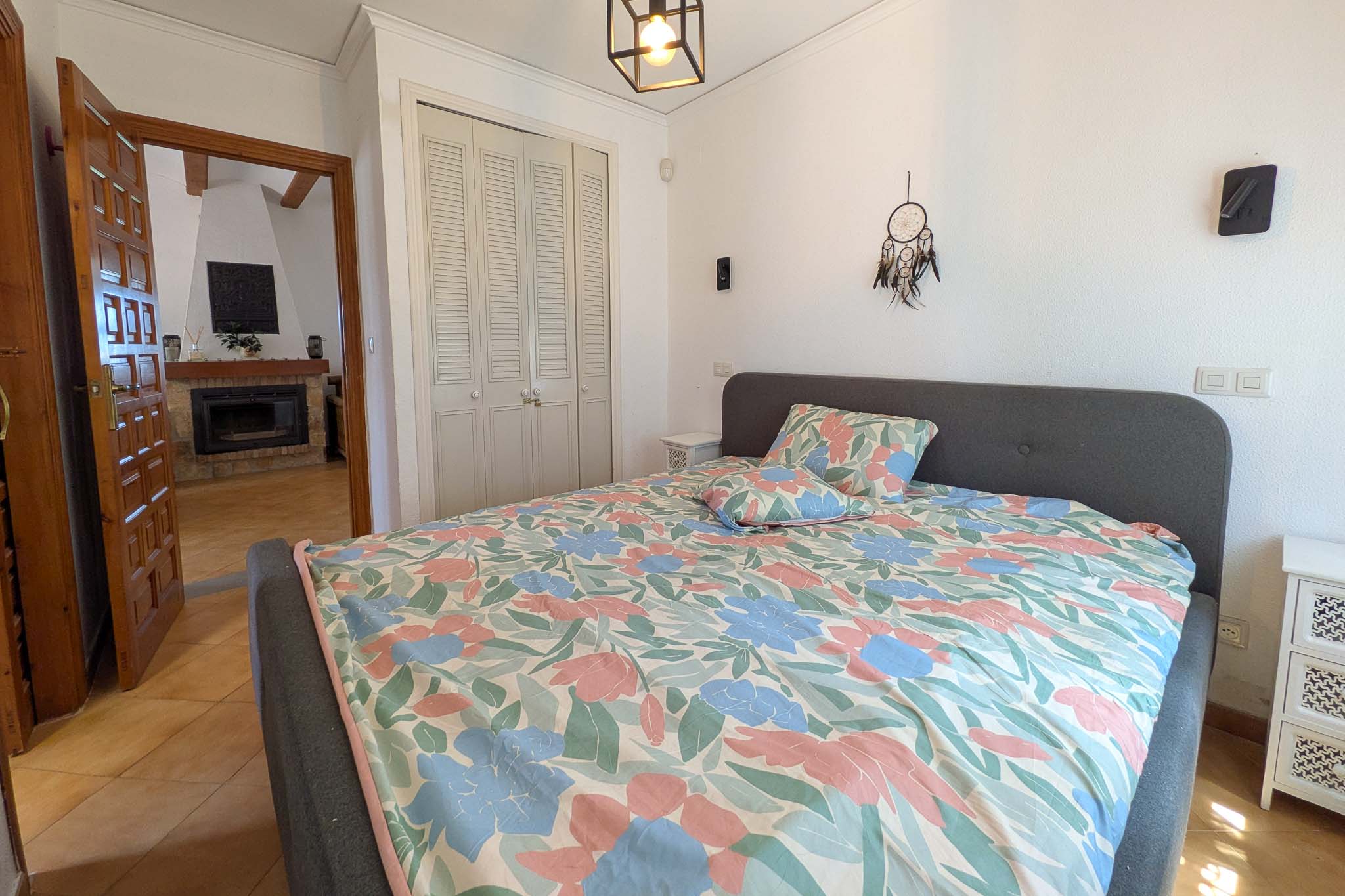 Lichte slaapkamer met ingebouwde kasten met witte lamellendeuren en een doorkijk naar de woonkamer met open haard via de houten deurpost.