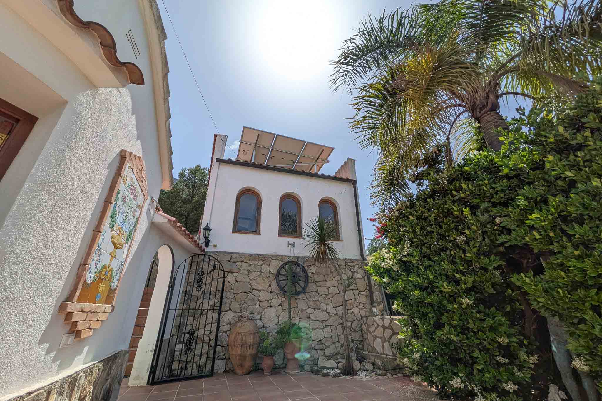 Mediterrane gevel met natuurstenen muren, zonnepanelen op het dak en traditionele details zoals een keramisch wandtableau en een smeedijzeren hek.