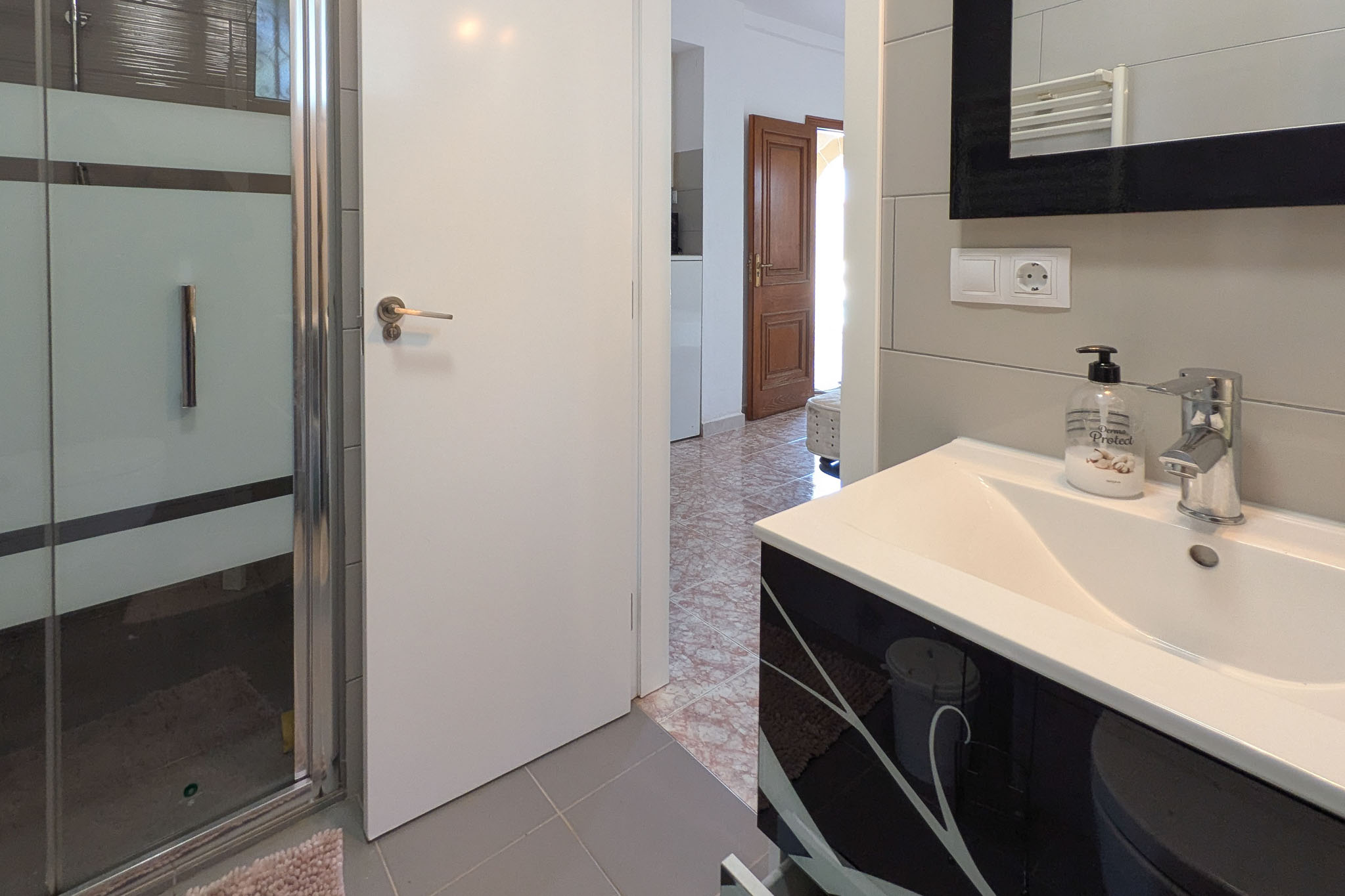 Moderne badkamer met een zwart gedecoreerd wastafelmeubel, glazen douchecabine en grijze betegeling. De witte deur biedt toegang tot een gang met traditionele vloerpatronen.