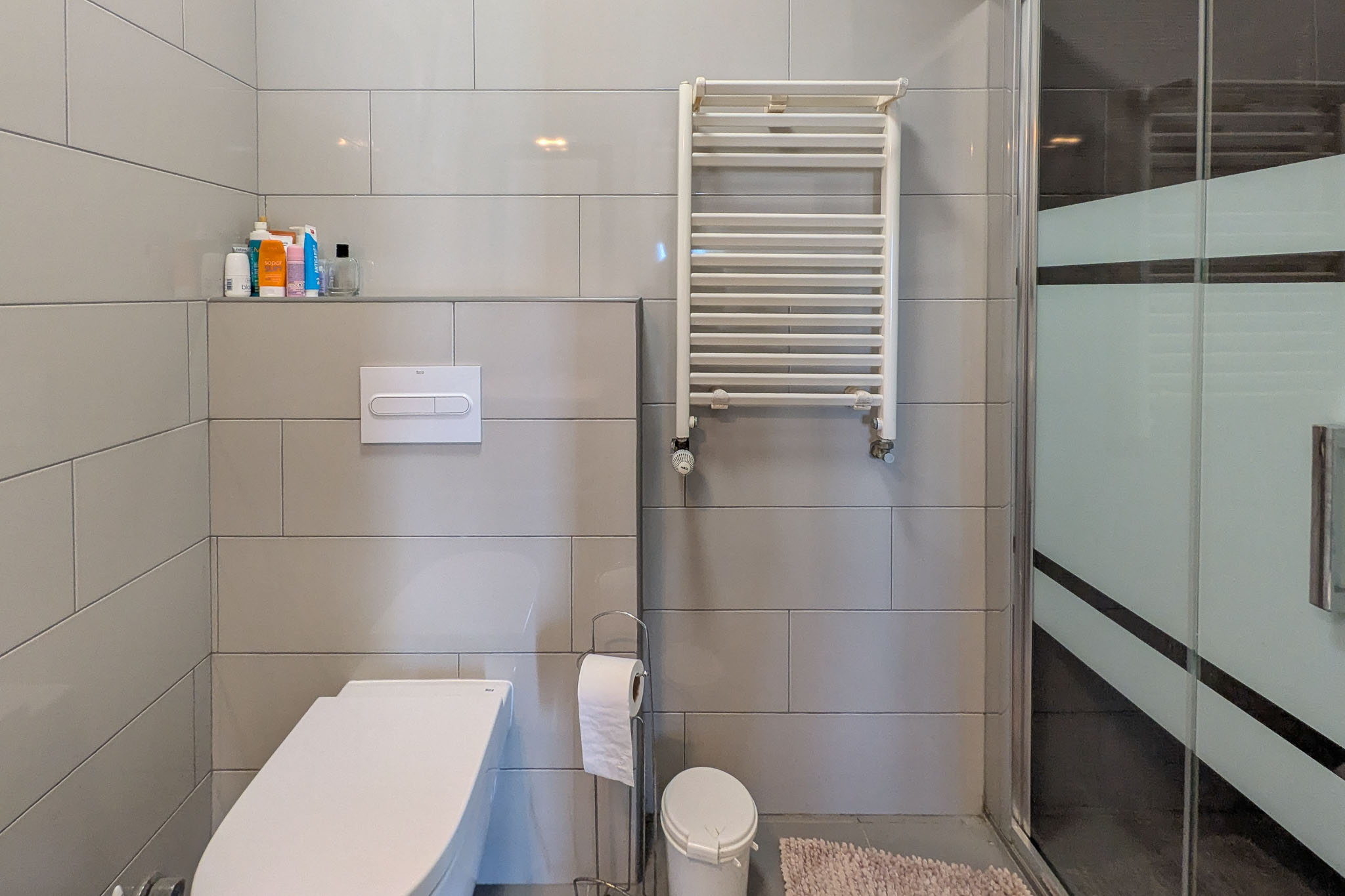Moderne badkamer met hangend toilet, grijze wandtegels, een witte handdoekradiator en een glazen douchewand met een matte privacyband.