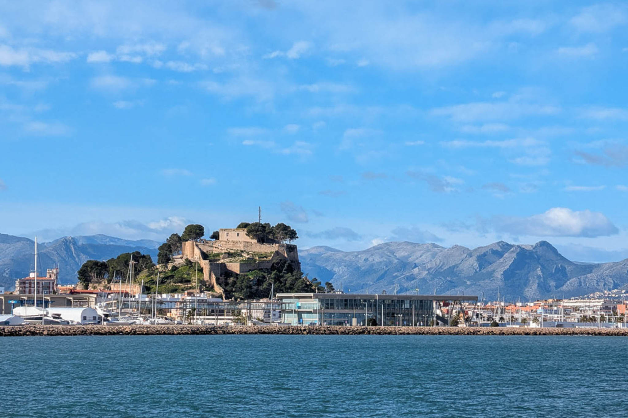 Panoramisch uitzicht op het kasteel van Dénia en de jachthaven, met de historische vesting op de heuvel en de bergen aan de horizon.