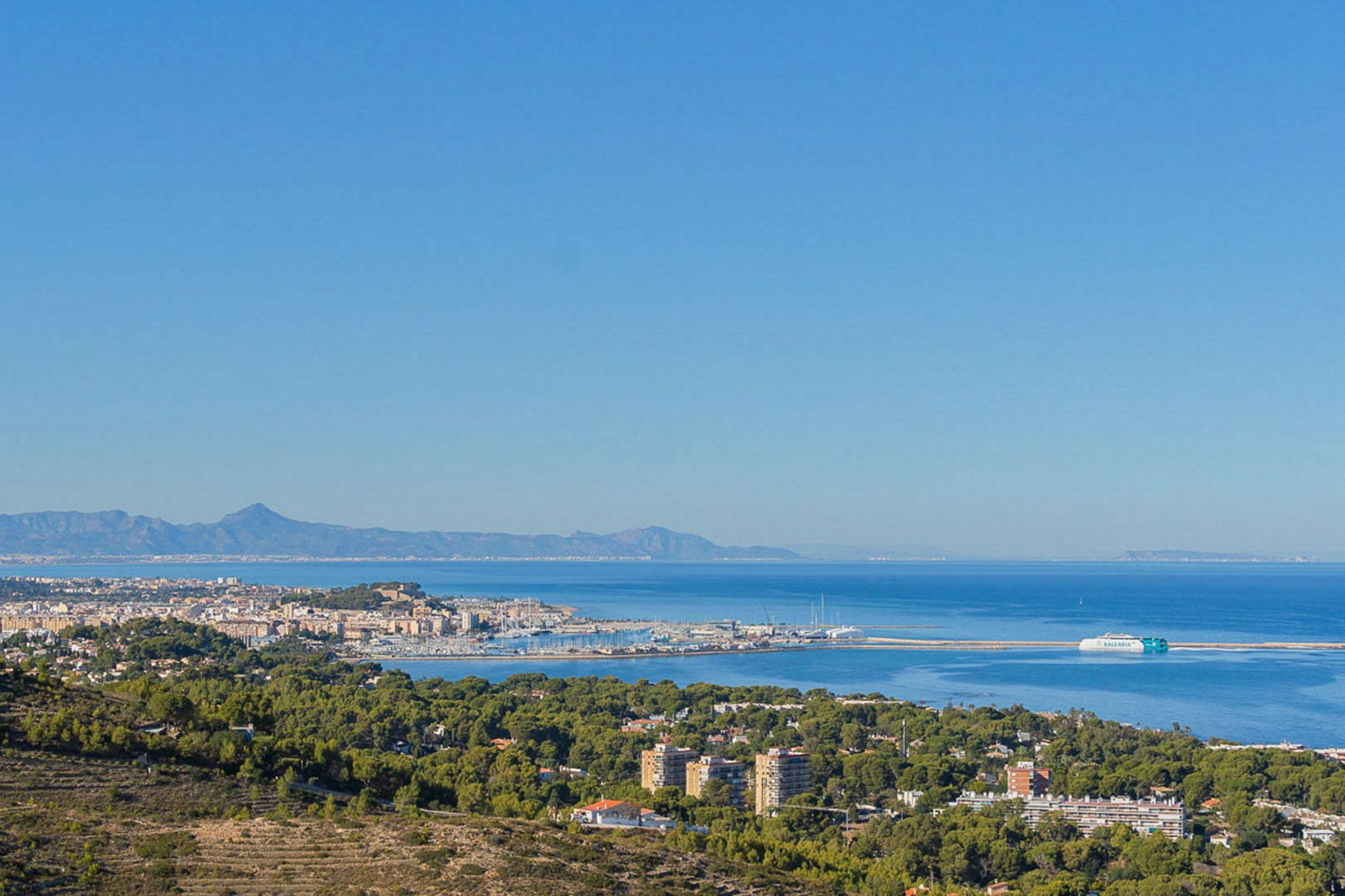 Panoramisch uitzicht vanaf de woning op de haven van Dénia en de Middellandse Zee, met de veerboot en de verre bergketens aan de horizon.