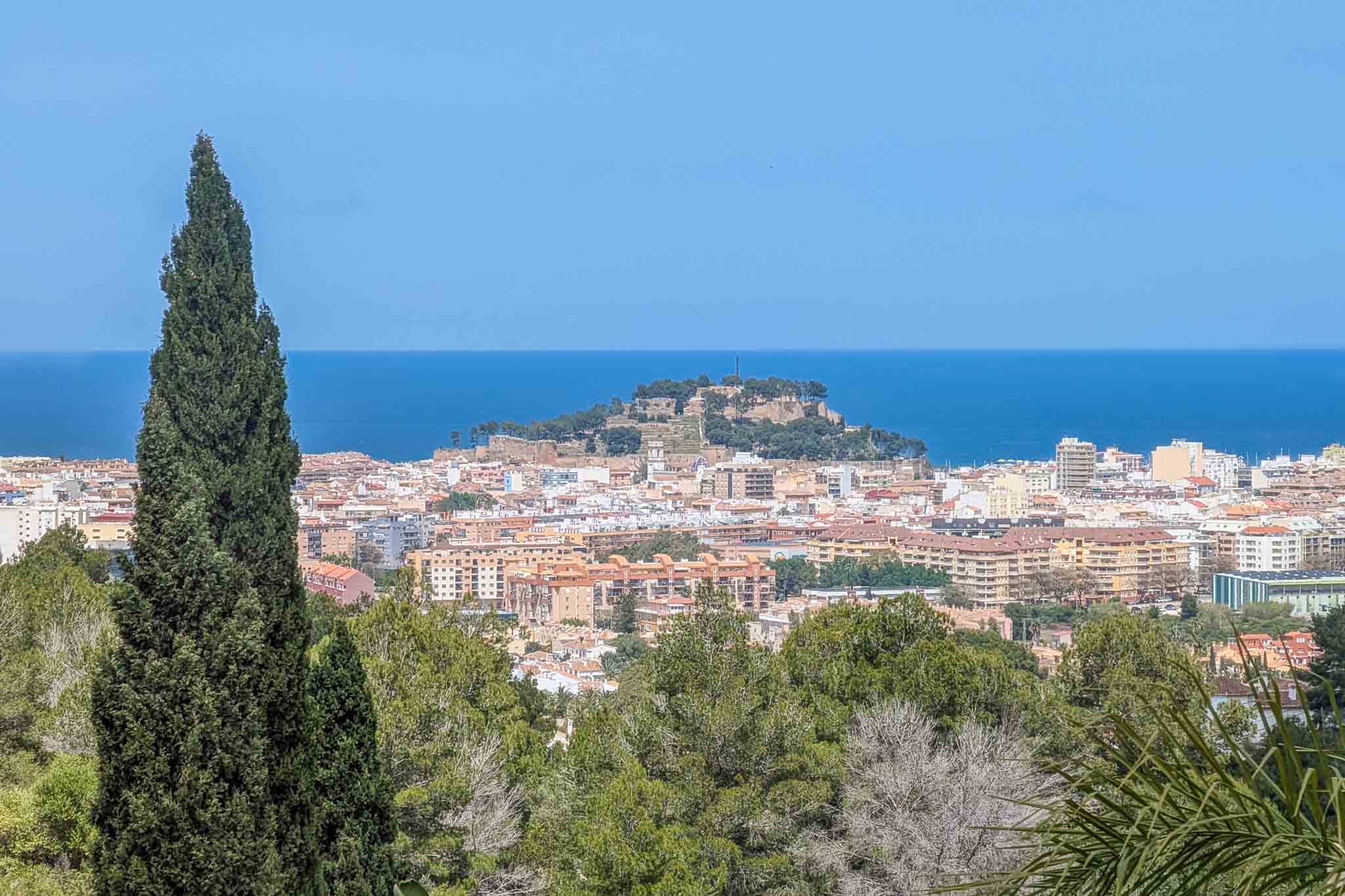 Panoramisch uitzicht vanaf het perceel op het kasteel van Dénia en de Middellandse Zee, omlijst door lokale vegetatie en cipressen.