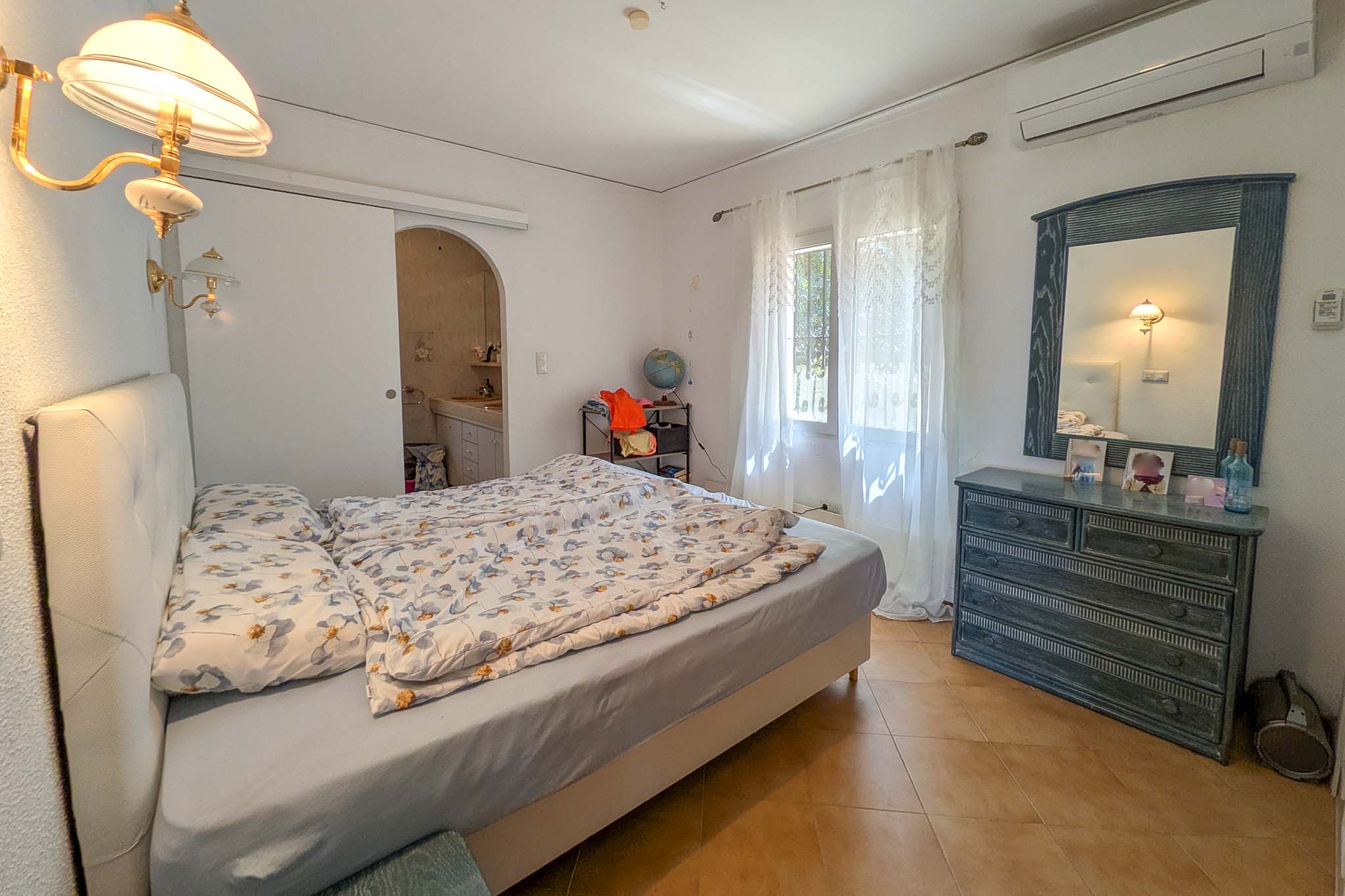 Slaapkamer met en-suite badkamer, split-airconditioning, terracotta vloertegels en houten meubels. De ruimte geniet van natuurlijk licht en een klassieke afwerking.