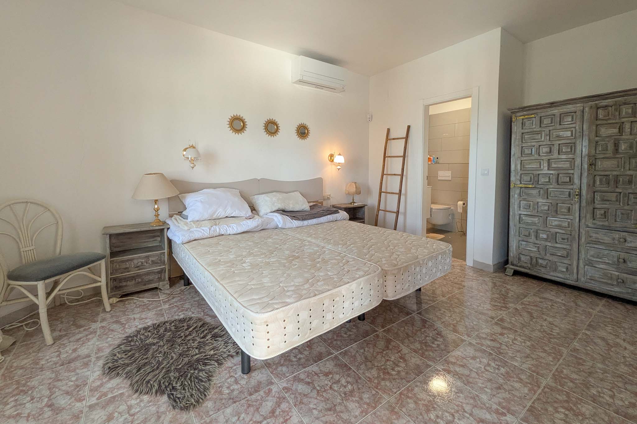 Slaapkamer met en-suite badkamer, tegelvloer met marmereffect, rustieke handgesneden houten kast en wandgemonteerde airconditioning.