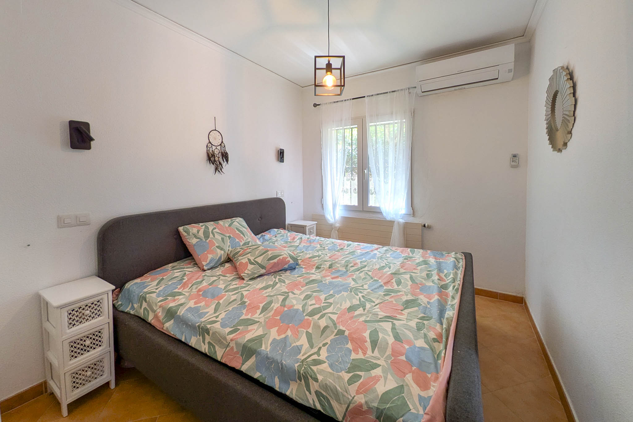 Slaapkamer met grijs gestoffeerd bed, split-airconditioning en een terracotta tegelvloer. De kamer heeft natuurlijke lichtinval en een industriële hanglamp.