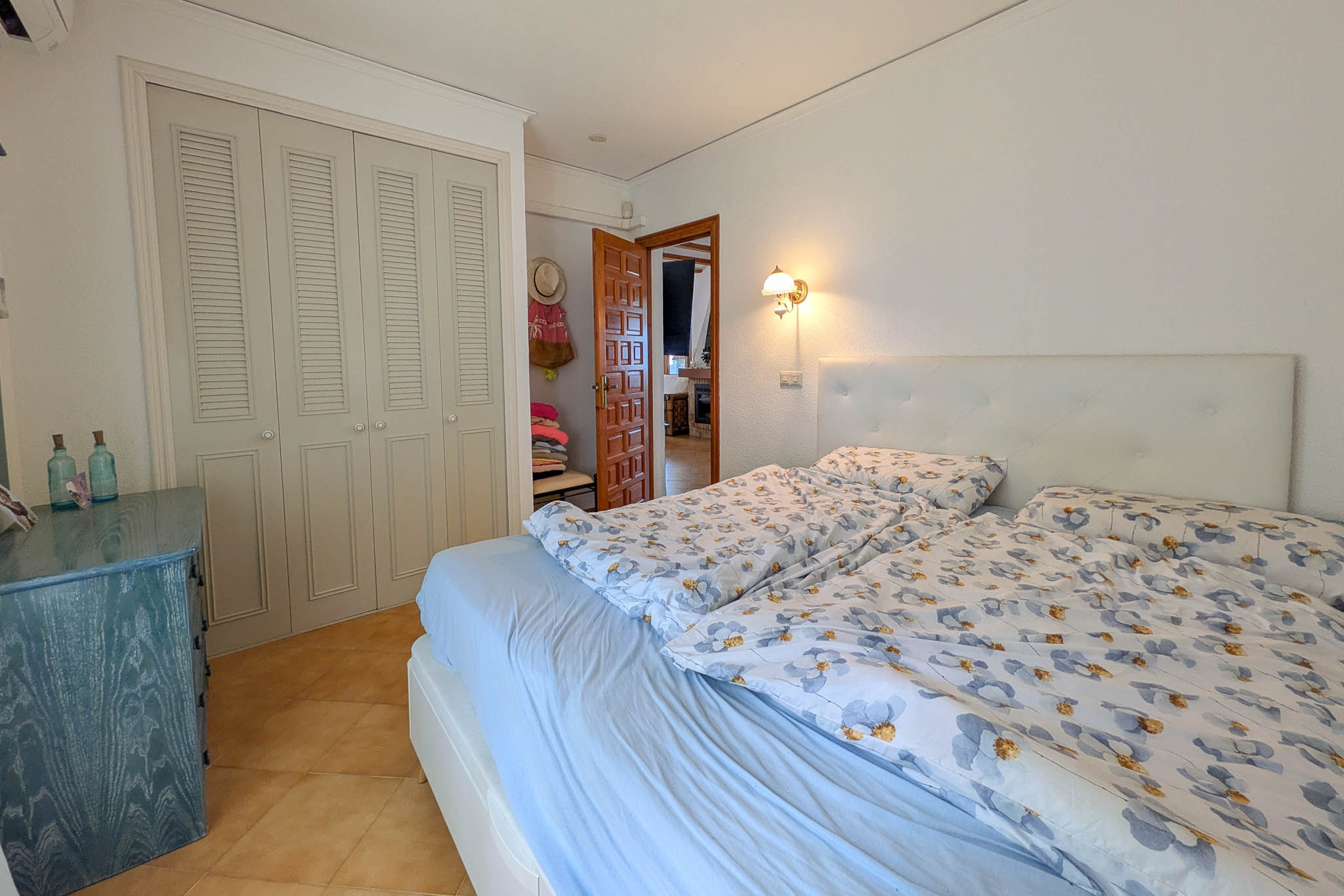 Slaapkamer met ingebouwde kasten met lamellendeuren, keramische tegelvloer en een gecapitonneerd hoofdbord. Een blauwe houten ladekast siert de lichte ruimte.