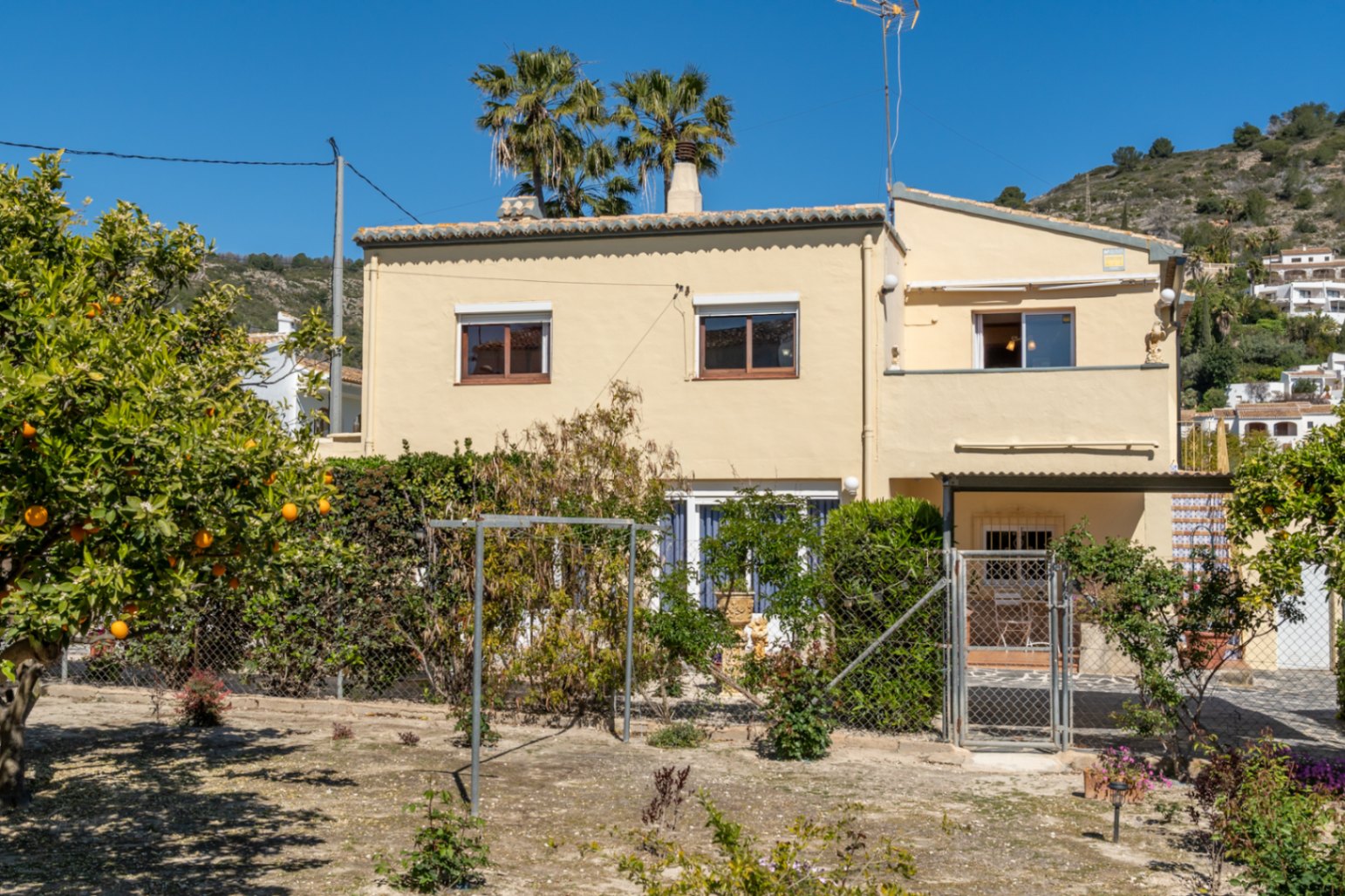 Buitengevel van de villa met crème muren, omringd door een tuin met sinaasappelbomen en palmen tegen een mediterraan heuvellandschap.