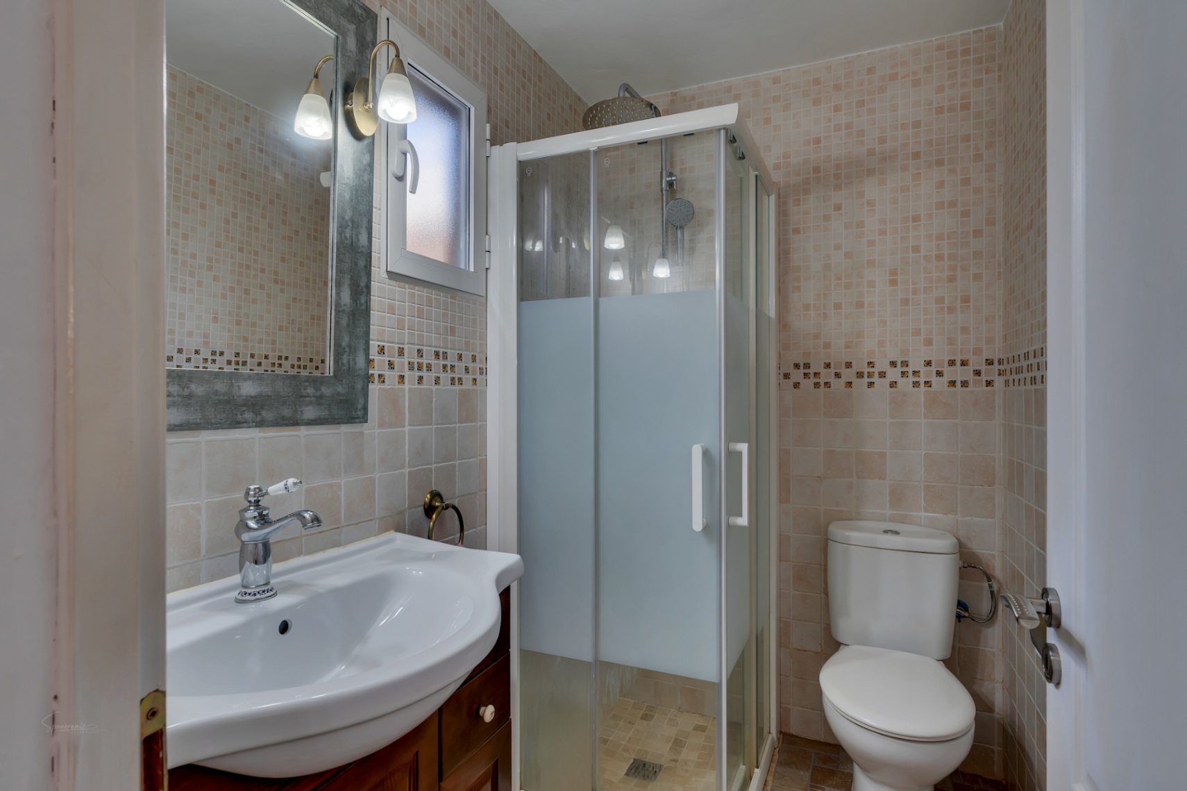 Functionele badkamer met beige wandtegels, glazen douchecabine, wastafel op houten meubel en een spiegel met klassieke wandverlichting.