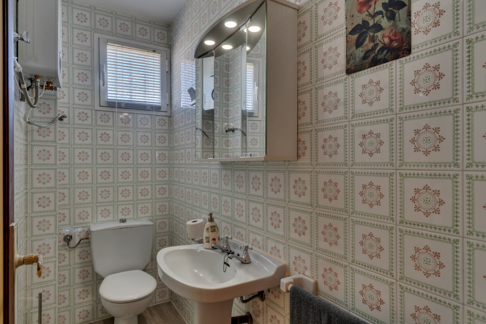 Functionele badkamer met gedecoreerde wandtegels, een wastafel op zuil, toilet en een spiegelkast met ingebouwde verlichting.