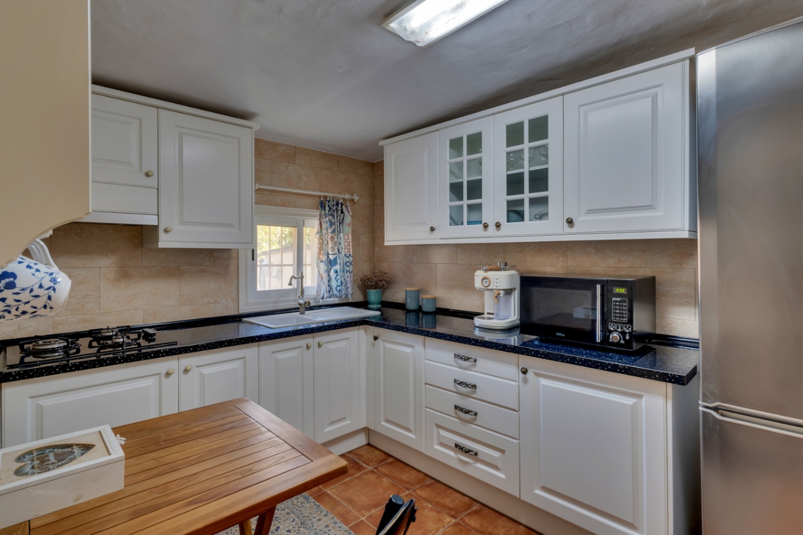 Functionele keuken met witte kasten, een donker granieten werkblad en rvs-apparatuur. Voorzien van een gaskookplaat, terracotta vloertegels en natuurlijke lichtinval.