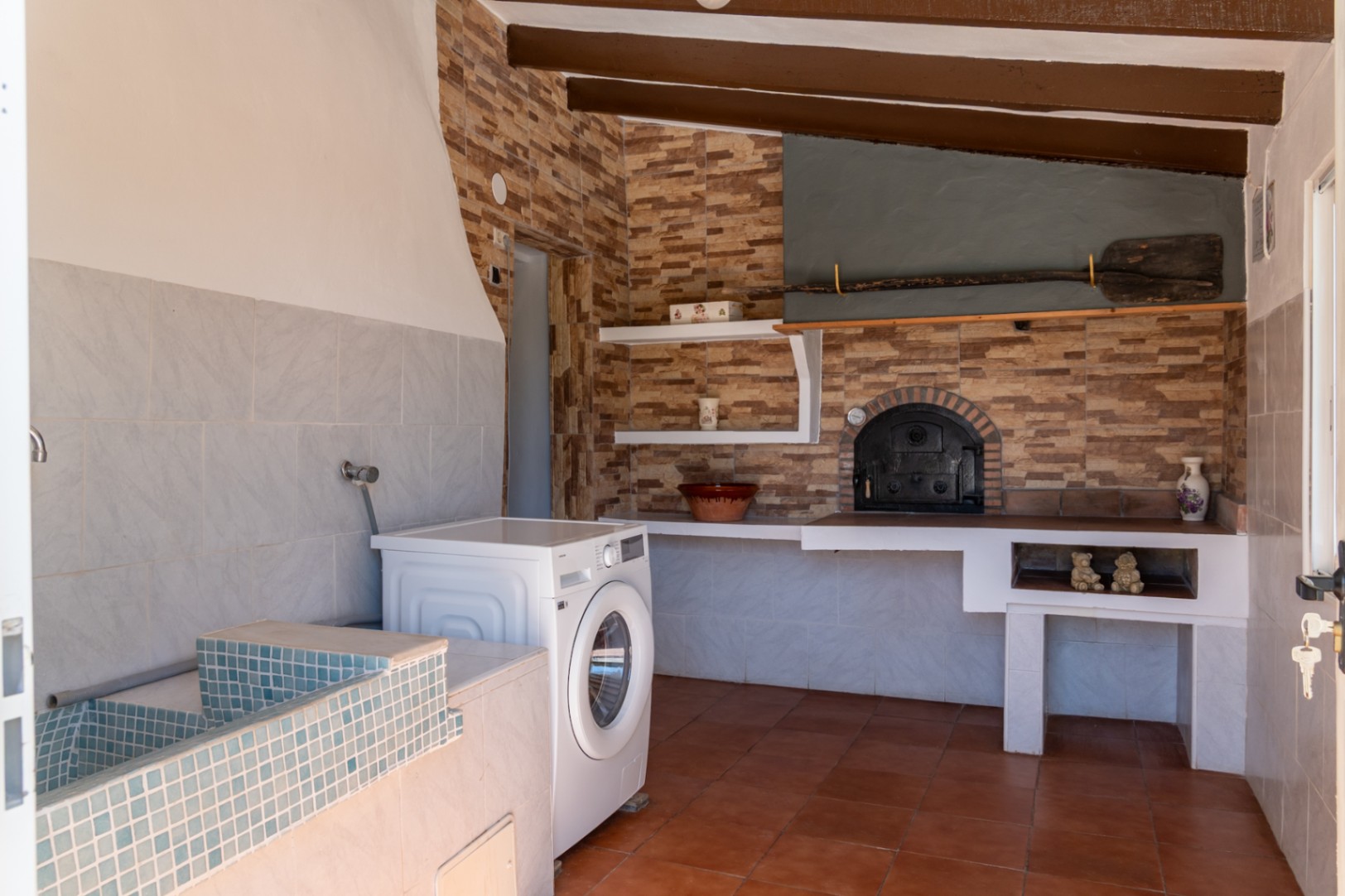 Functionele wasruimte en rustieke keuken met blauwe mozaïek wasbak, wasmachine en een traditionele houtoven onder donkere houten balken en terracotta tegels.
