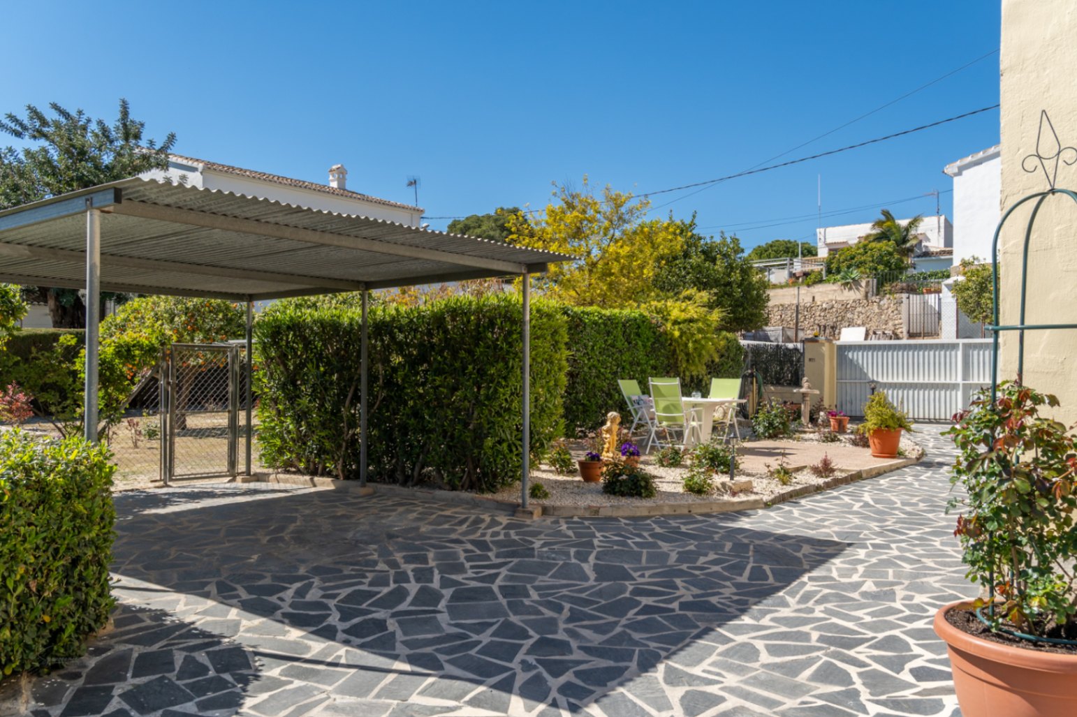 Ingang van de woning met natuurstenen bestrating, een metalen carport en een aangelegde tuin met hagen en een gezellig terrasgedeelte.