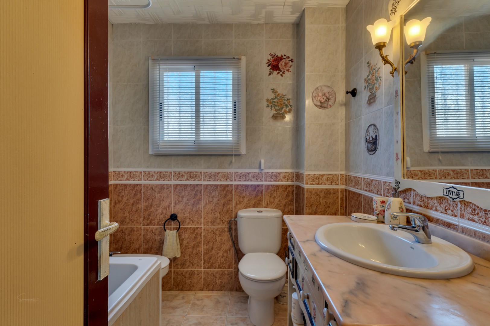 Klassieke badkamer met marmeren wastafelblad, wandtegels met bloemmotieven, wit sanitair en sfeervolle vintage wandverlichting.