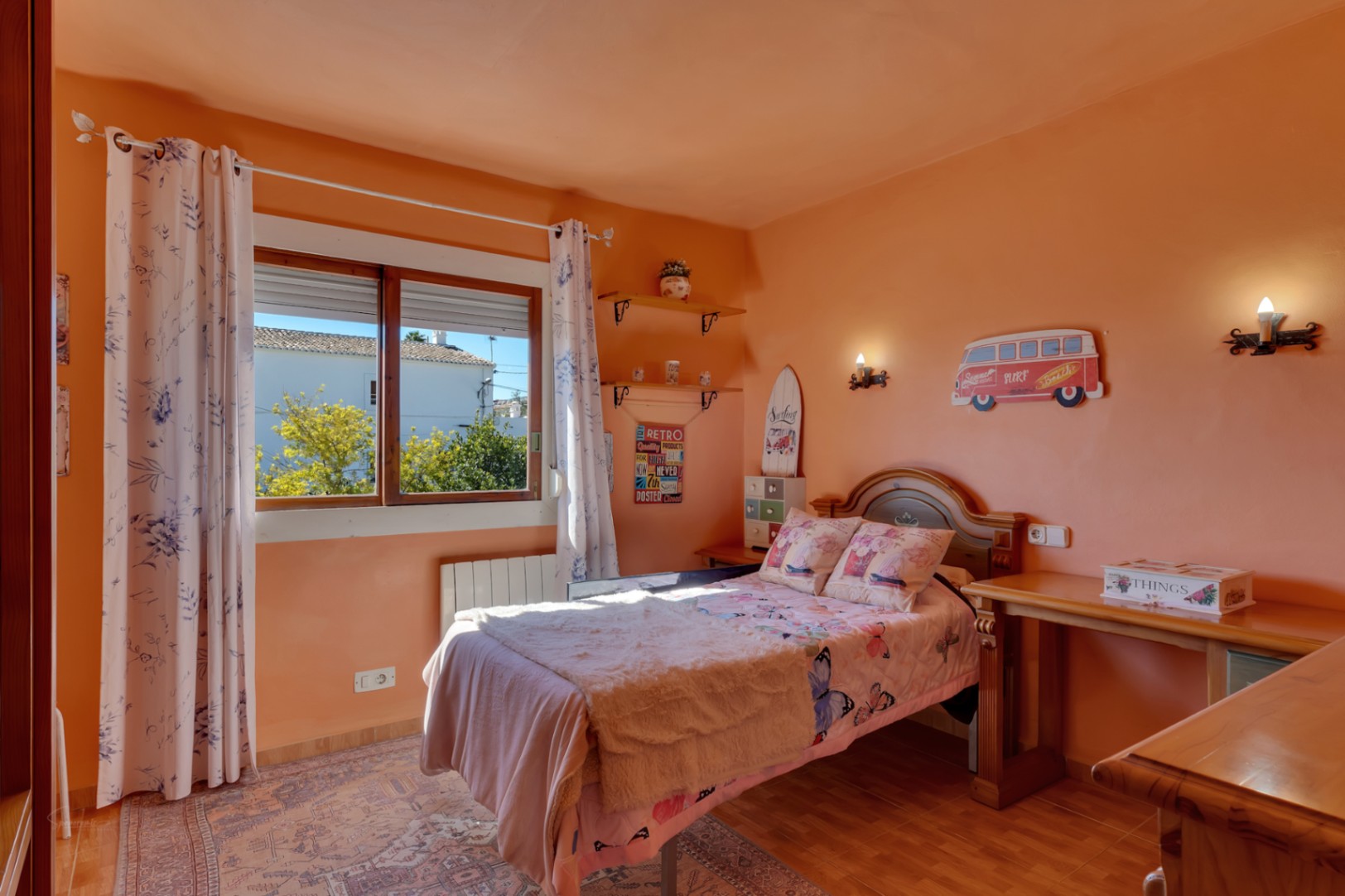 Lichte slaapkamer met zalmkleurige muren, eenpersoonsbed, houten bureau en raam met uitzicht naar buiten. Voorzien van radiator en een tegelvloer.