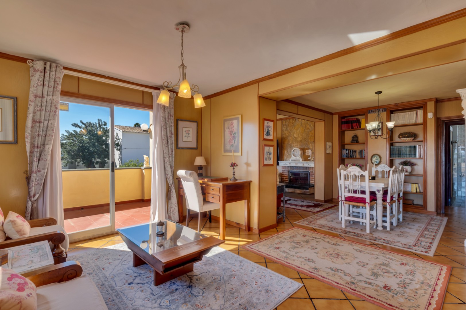 Lichte woon- en eetkamer met houten bureau, tegelvloer en toegang tot het terras. Op de achtergrond een eethoek met ingebouwde boekenkast en open haard.