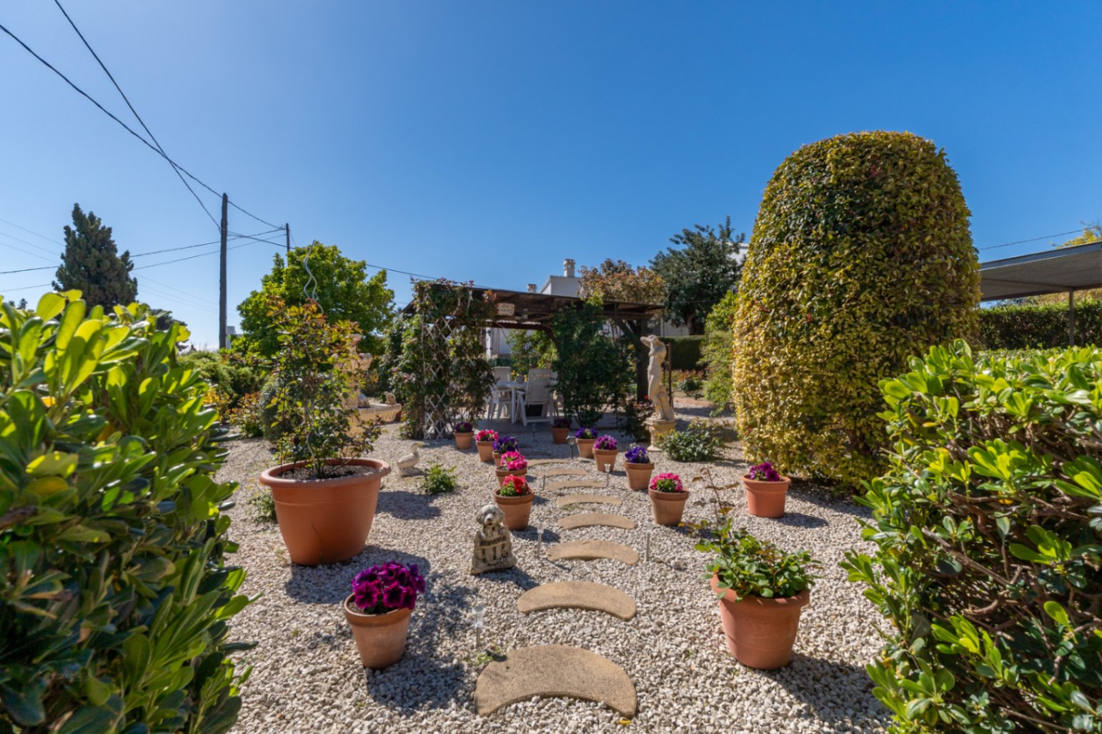 Mediterrane tuin met wit grind, een stenen pad en terracotta potten. Een houten pergola herbergt een eethoek buiten, omgeven door strak gesnoeide hagen.