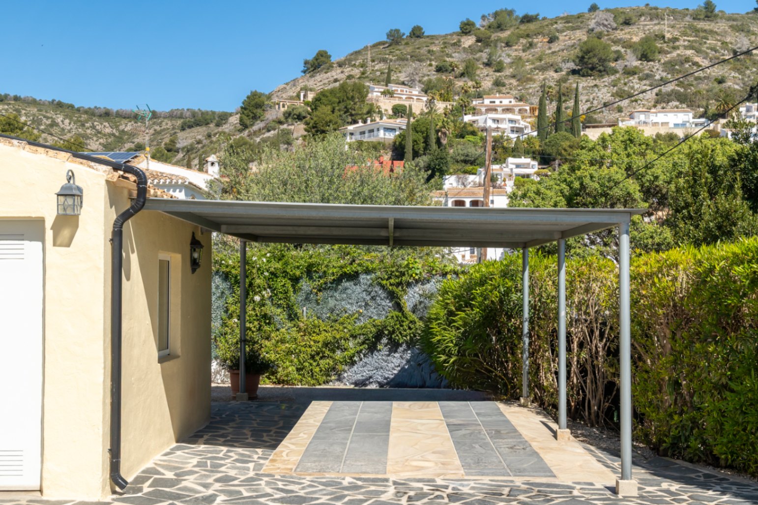 Metalen carport met plat dak en natuurstenen bestrating naast de villa, met uitzicht op de groene heuvels van Jávea.