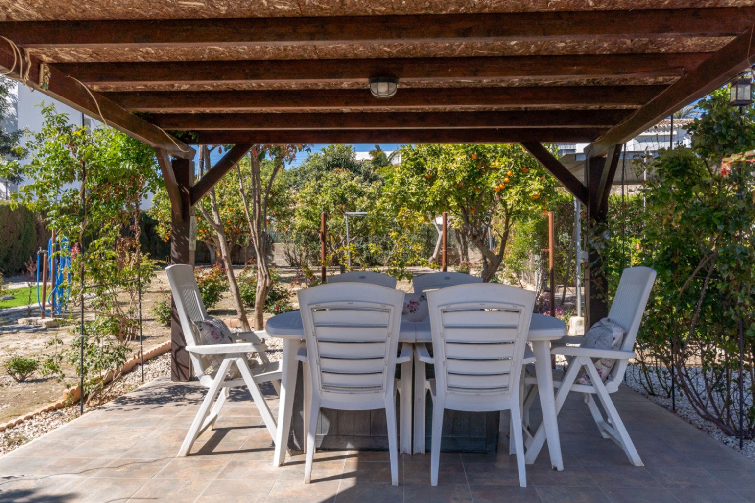 Overdekt buitenterras met een houten pergola, tegelvloer en een eethoek die uitkijkt op de tuin met fruitbomen.