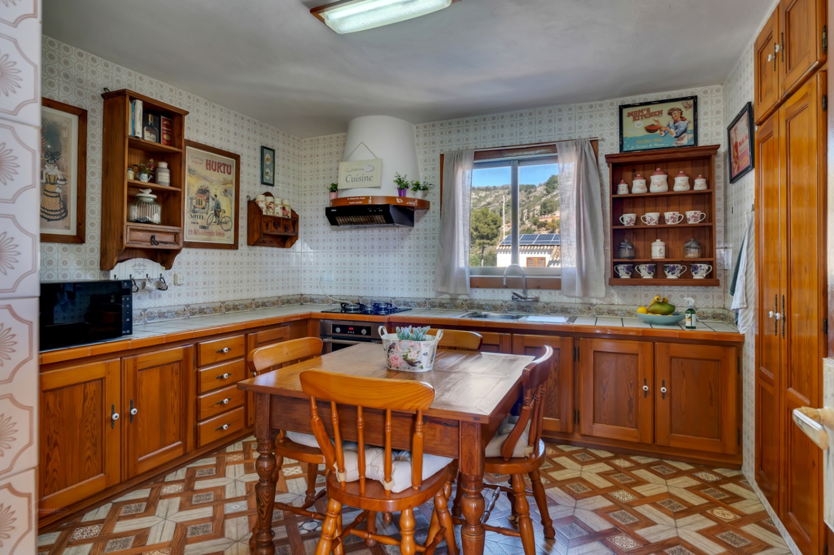 Rustieke keuken met houten kasten, een centrale eettafel en gedecoreerde wandtegels. Een raam biedt uitzicht op de omliggende heuvels en zorgt voor natuurlijk licht.