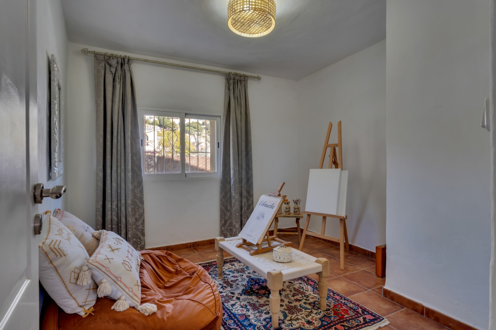 Veelzijdige kamer met terracotta vloertegels, ingericht als kunstatelier met houten ezel, natuurlijk licht en een sfeervol oosters tapijt.