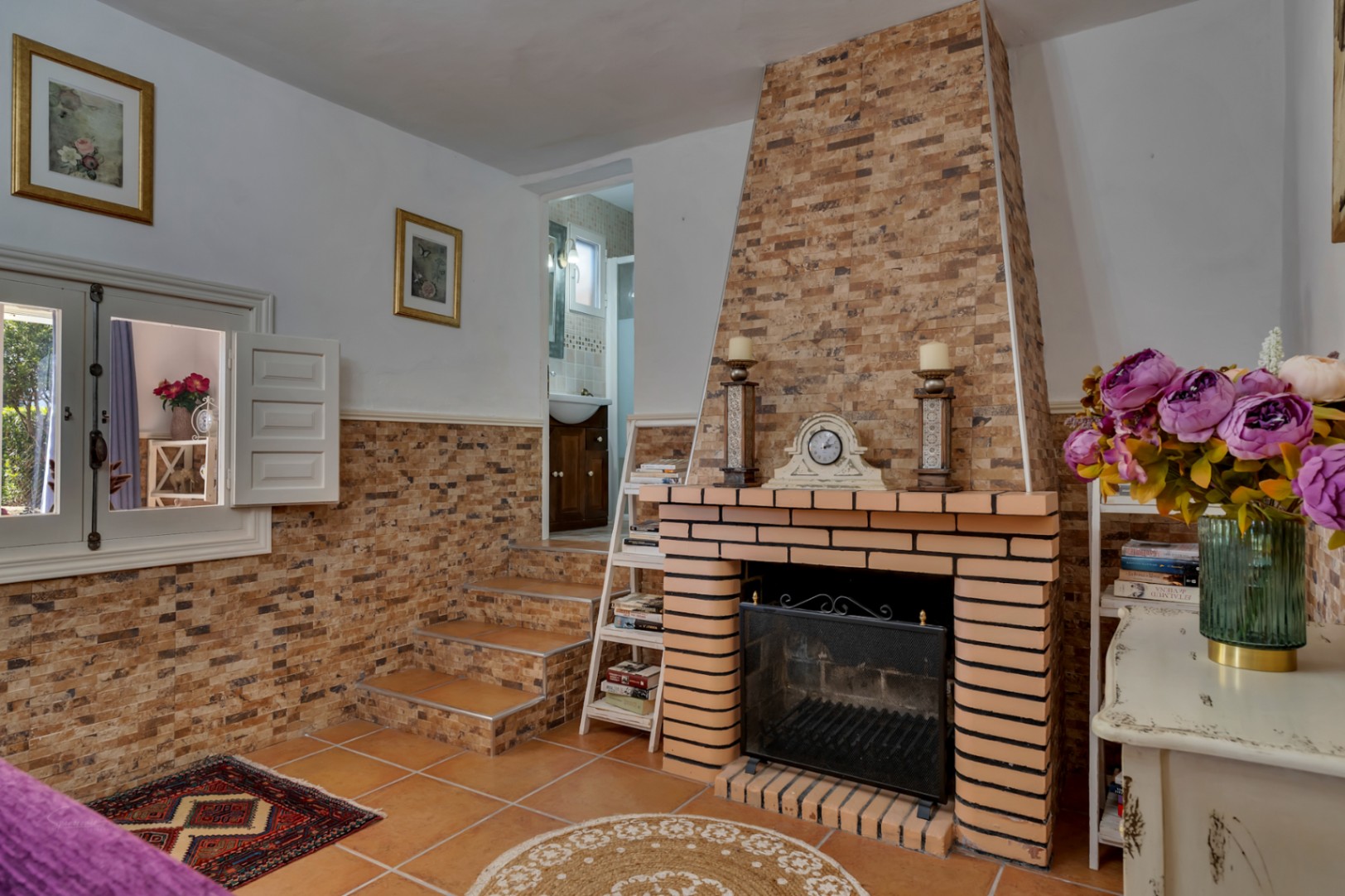 Woonkamer met een bakstenen open haard, terracotta vloertegels en een trappetje naar de badkamer. De ruimte is voorzien van authentieke witte binnenluiken en rustieke elementen.
