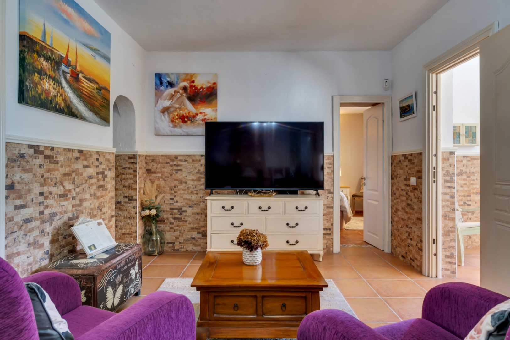 Woonkamer met paarse fauteuils, stenen wandbekleding, terracotta vloer en een wit tv-meubel met doorgang naar de slaapkamer.