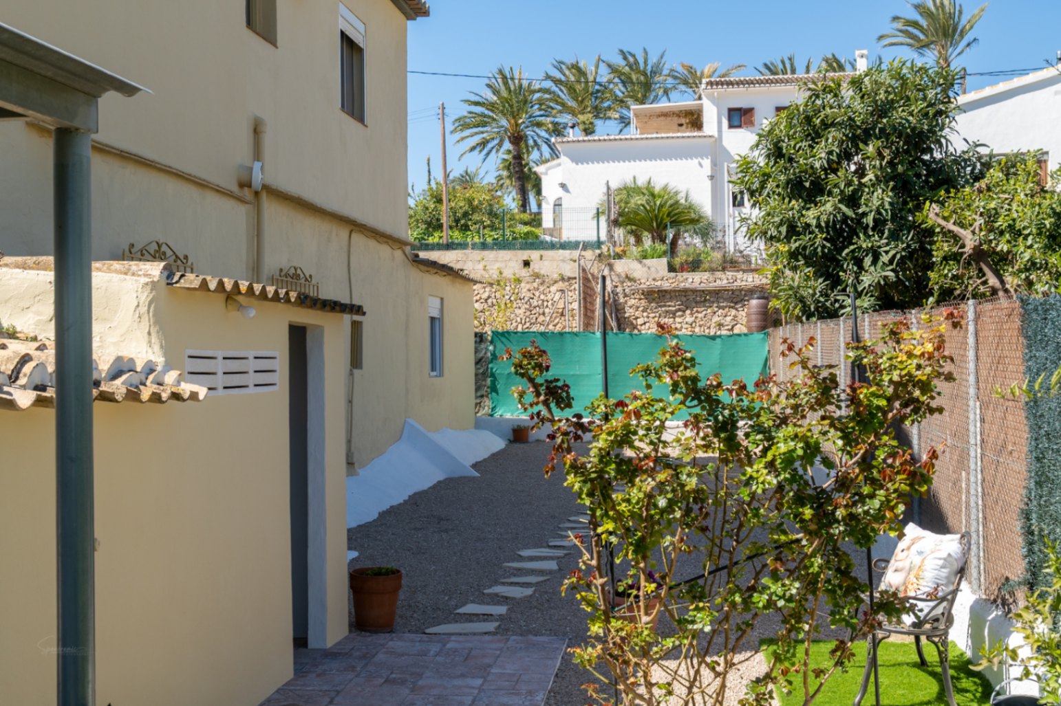 Zijtuin met grindpad, stapstenen, mediterrane beplanting en een functioneel bijgebouw op een zonnig perceel in Jávea.