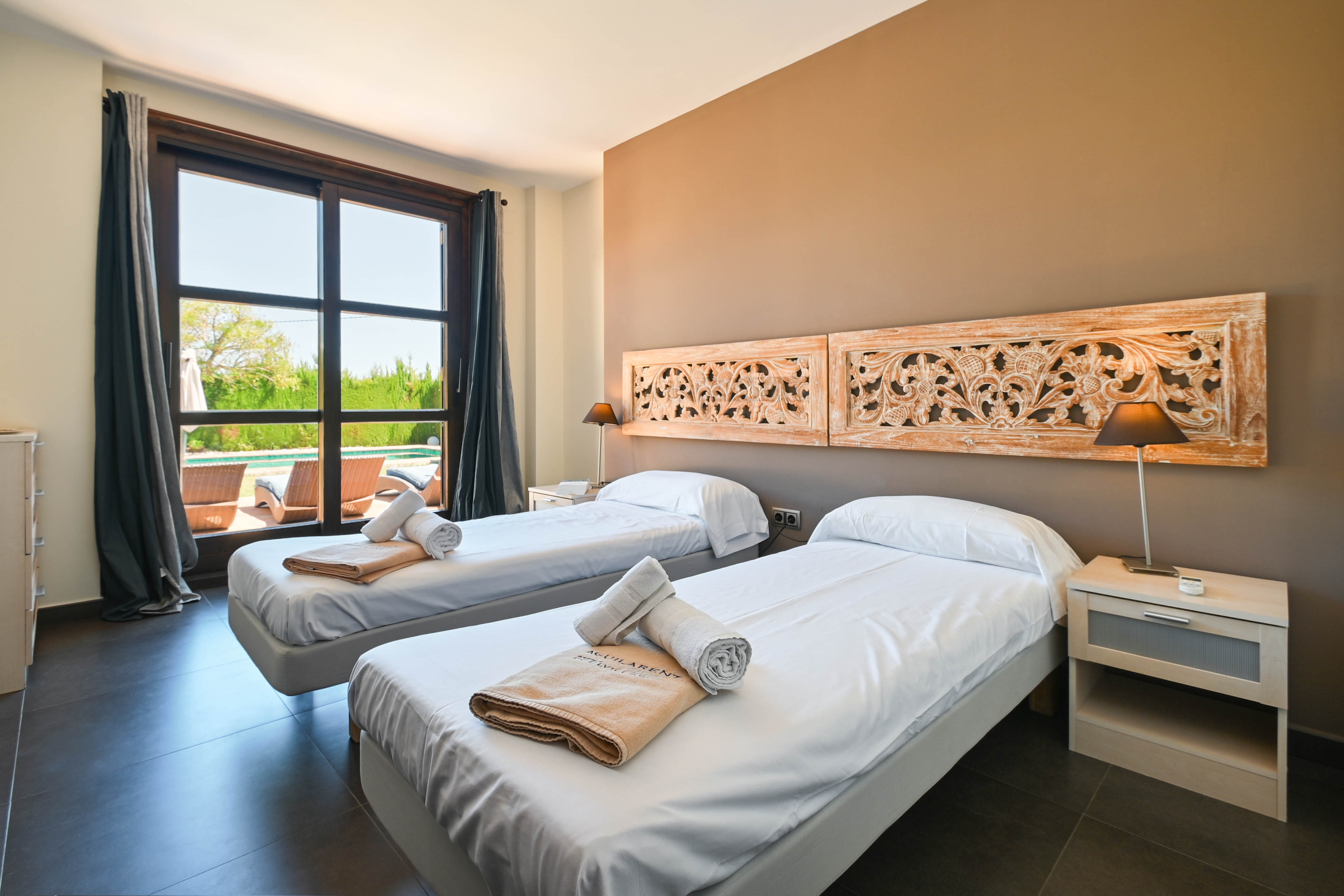Lichte slaapkamer met twee eenpersoonsbedden, gesneden houten hoofdeinden en uitzicht op het zwembad.