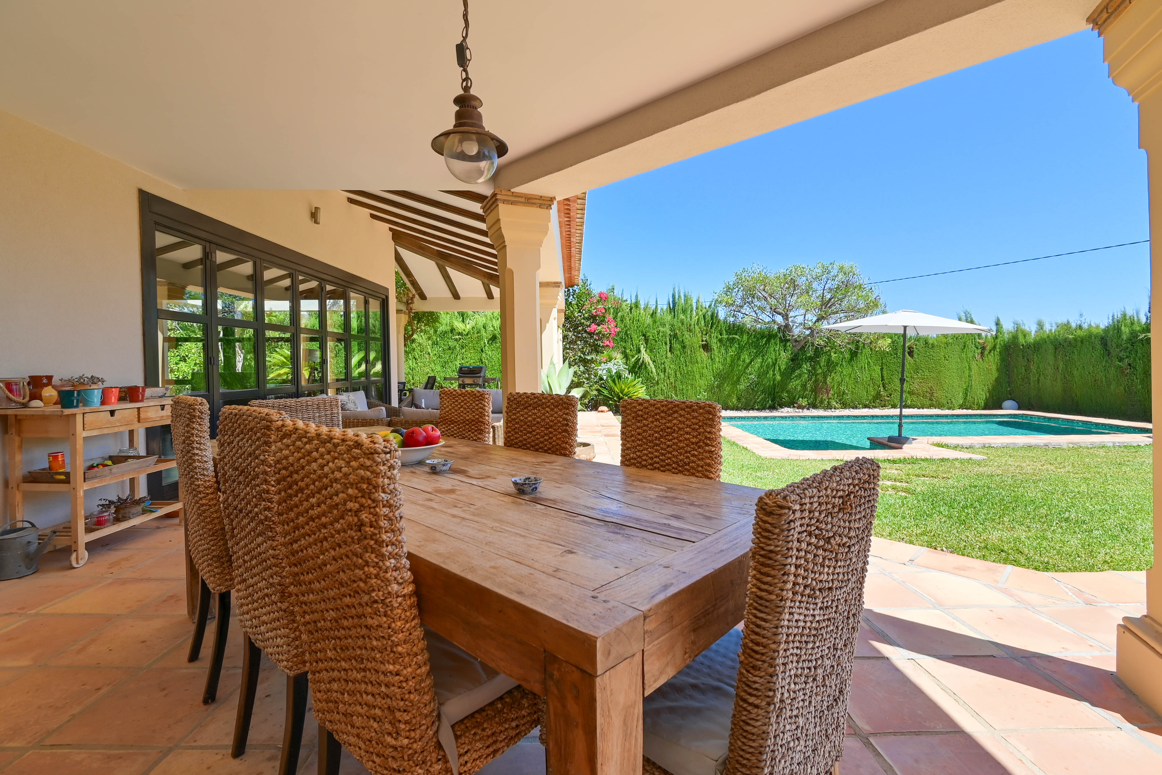 Overdekte eethoek buiten met massief houten tafel en rotan stoelen met uitzicht op het zwembad en de tuin.