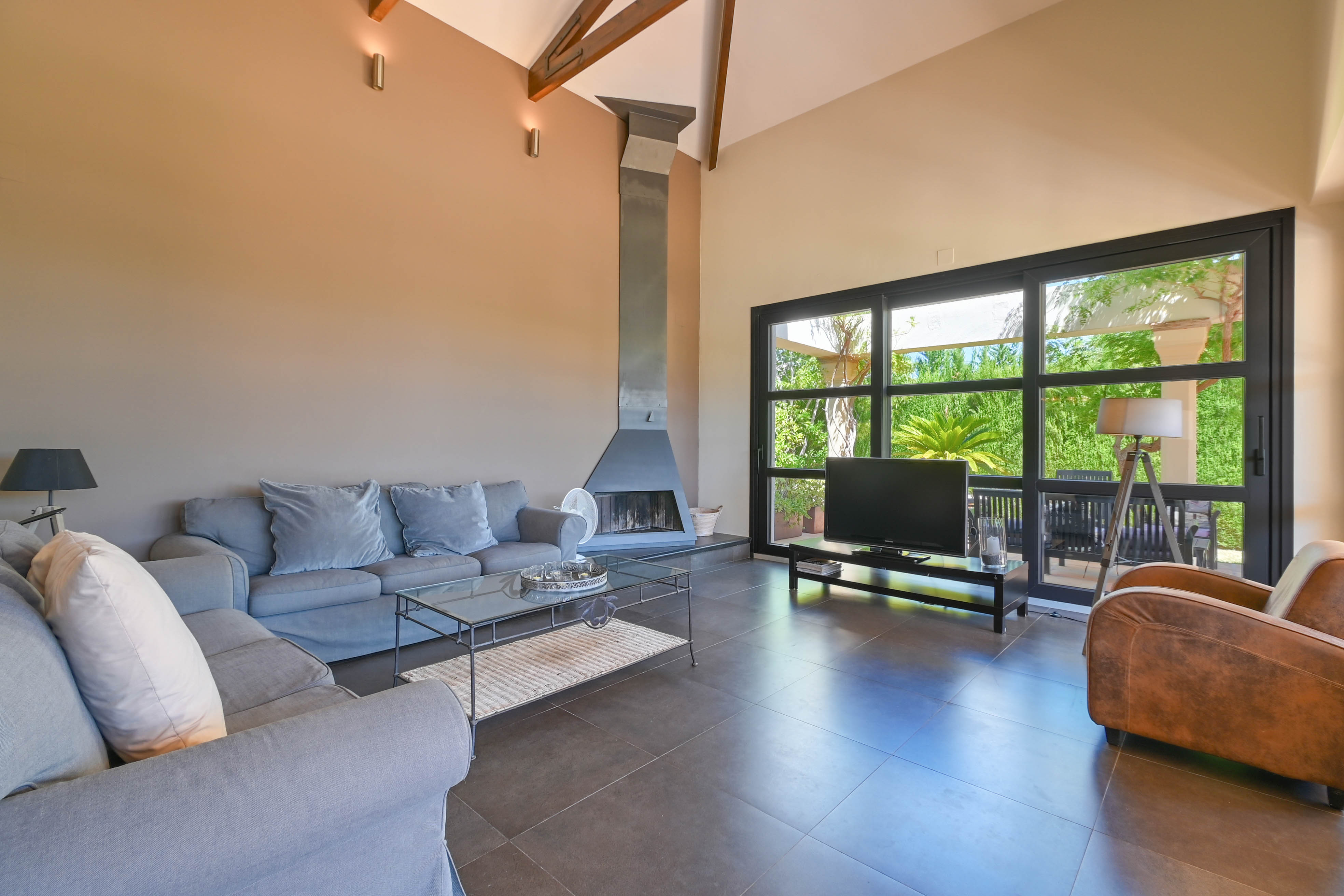 Ruime woonkamer met metalen open haard, grijze bank en grote ramen met uitzicht op de tuin in Denia, Costa Blanca.