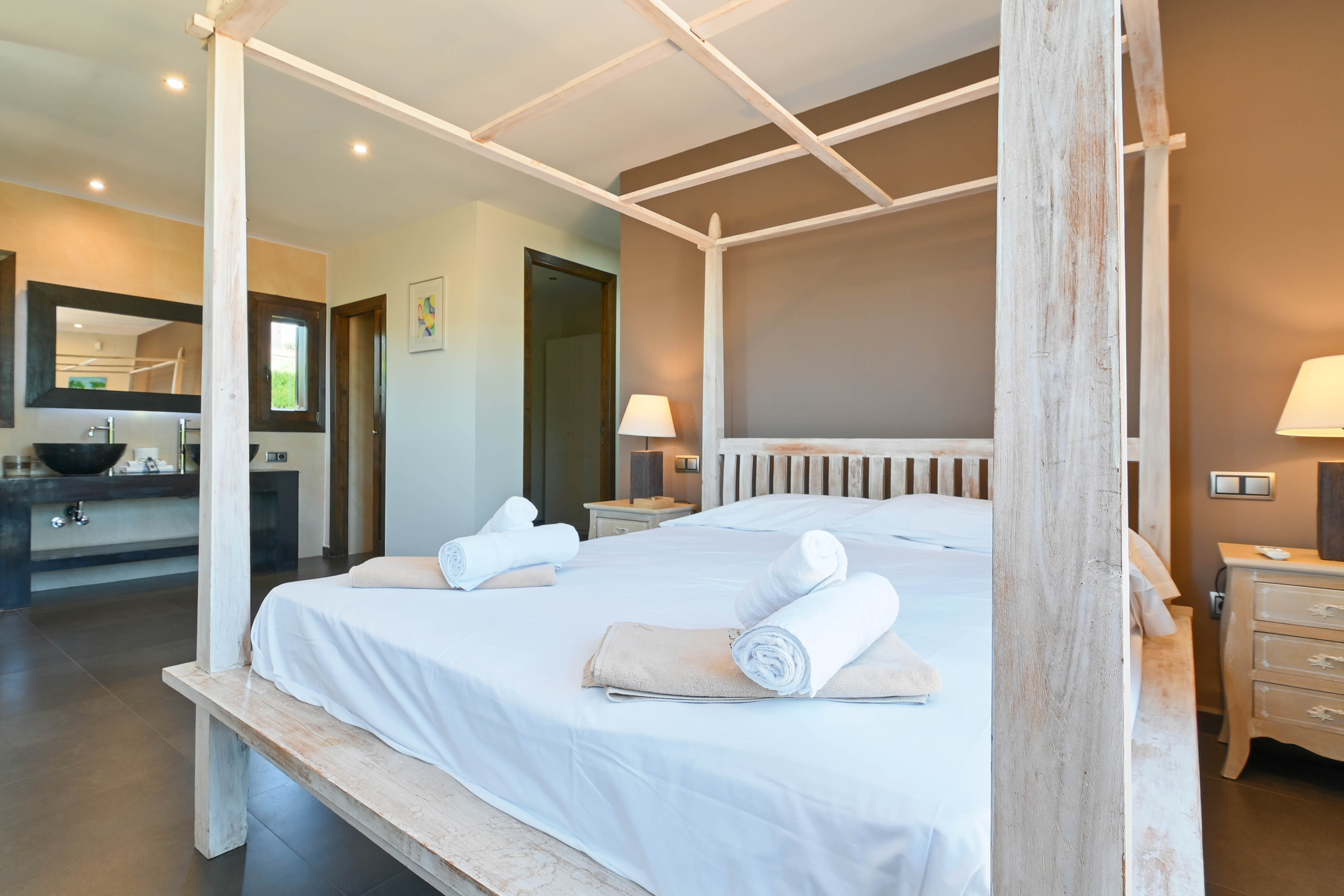 Slaapkamer met wit houten hemelbed, opgerolde handdoeken en badkamer met moderne wastafel.