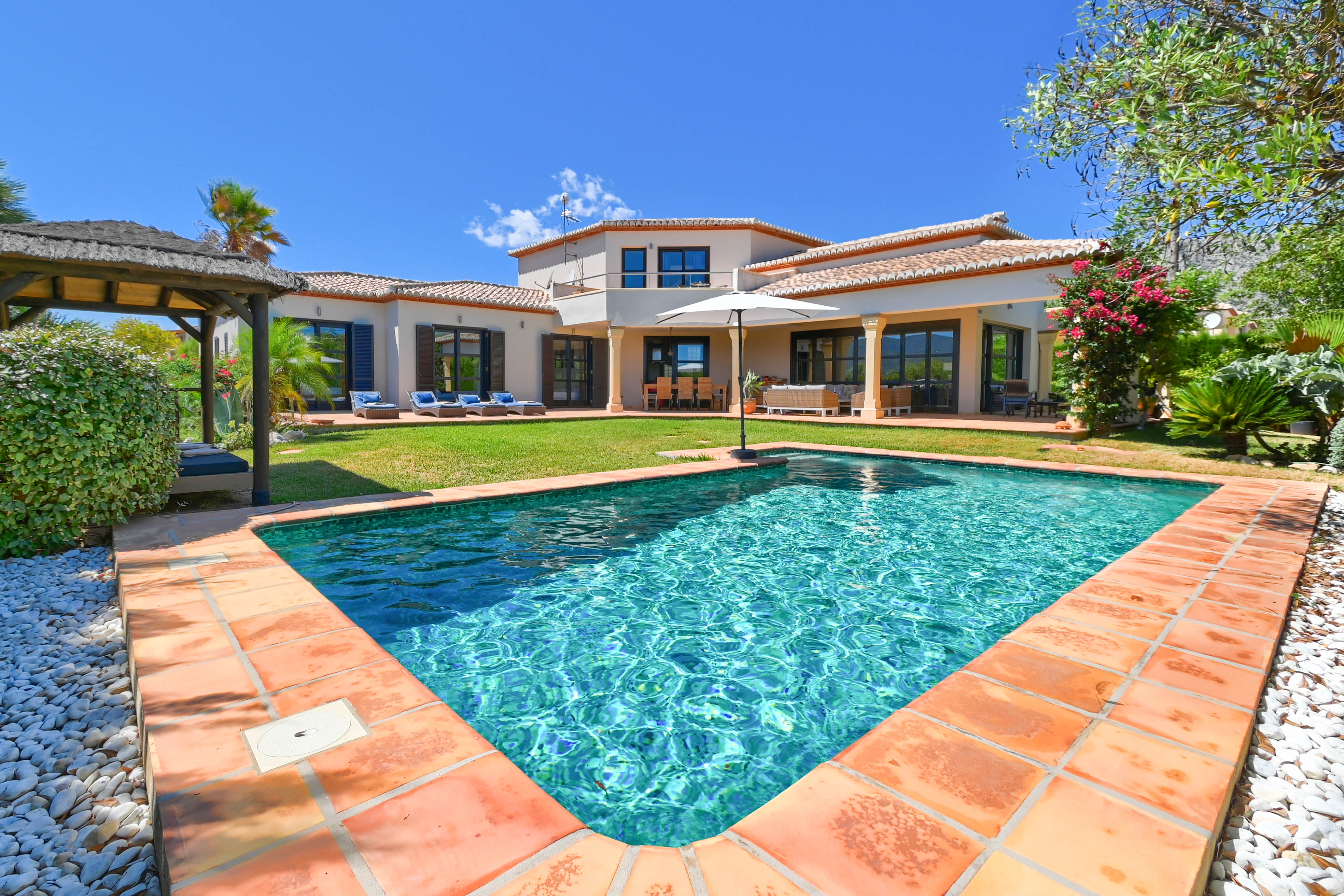 Uitzicht op het turquoise zwembad met terracotta rand en de villa met tuin en tuinhuis.