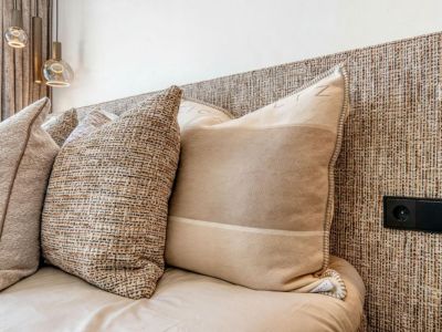 Detail van het bed met een geweven hoofdeinde en decoratieve kussens in neutrale tinten. Stopcontact geïntegreerd in het hoofdeinde.