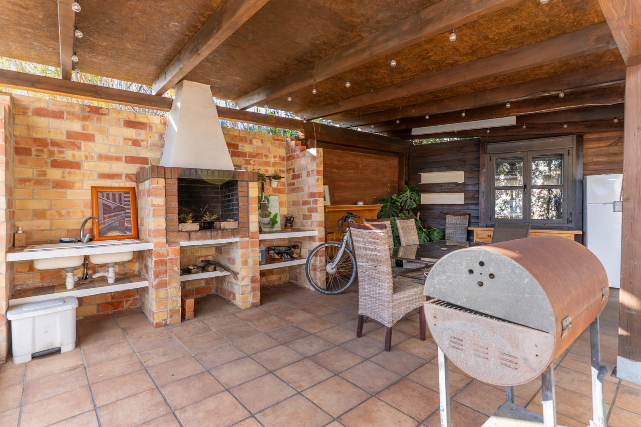 Buitenbarbecue met bakstenen oven, dubbele spoelbak en eethoek onder een houten dak met sfeerverlichting.