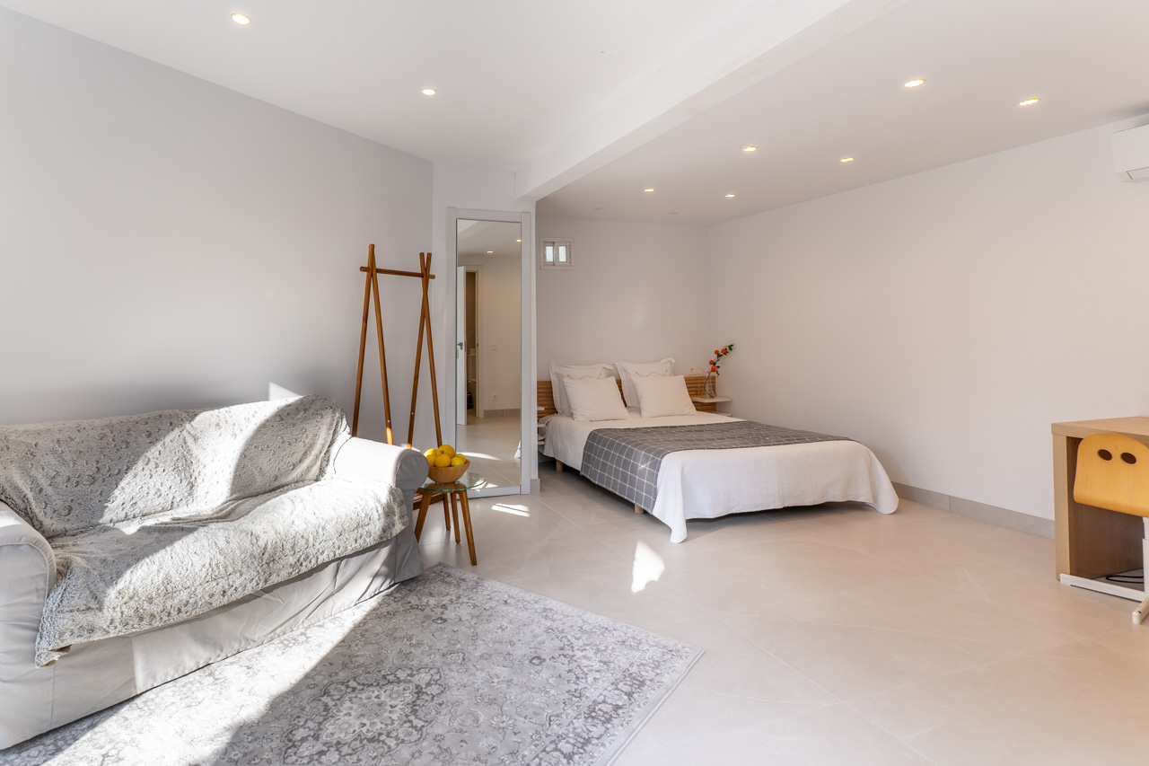 Lichte slaapkamer met sofa, tweepersoonsbed en bureau, verlicht door inbouwspots.