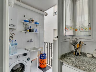 Wasruimte met wasmachine, gootsteen en witte betegelde planken. Raam met gordijnen met print.