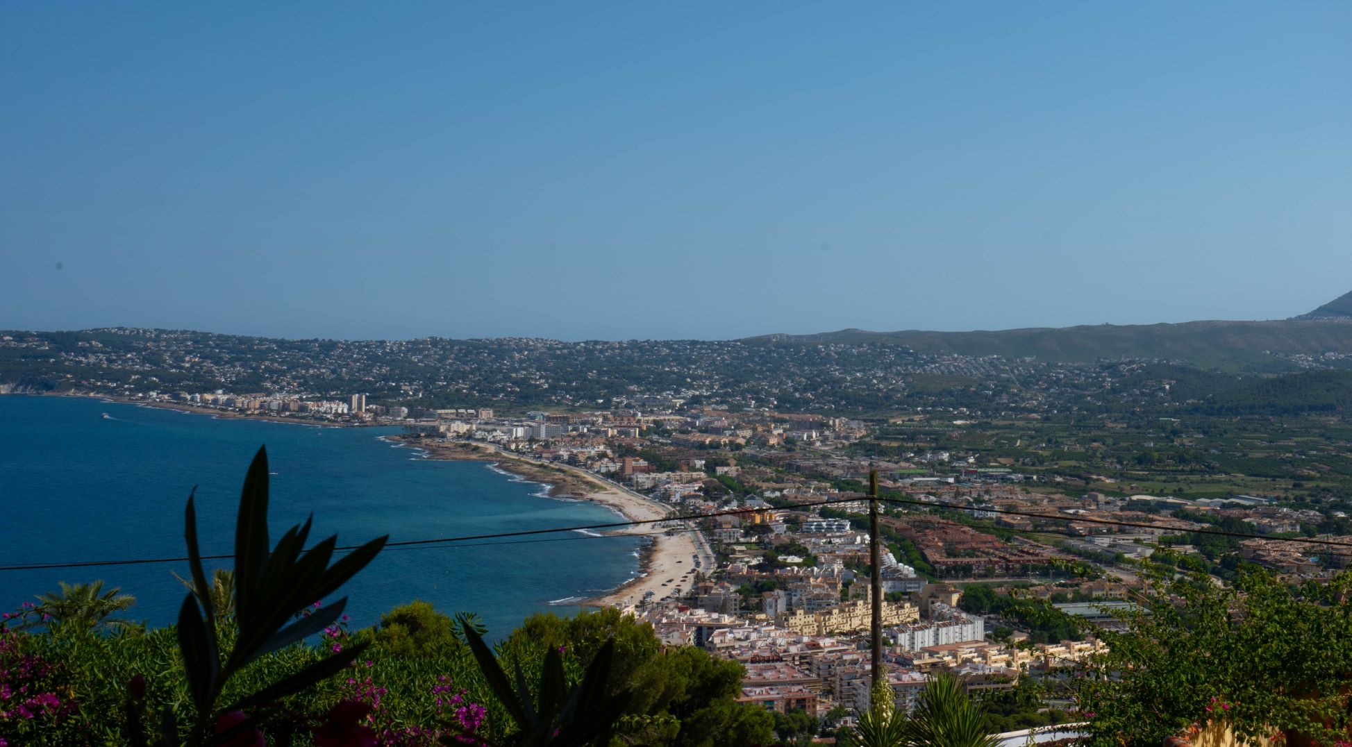 Panoramisch uitzicht op de baai van Jávea vanaf het perceel, met vegetatie op de voorgrond.