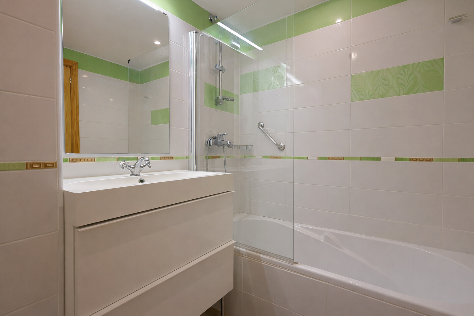 Badkamer met moderne wastafel, witte tegels met groene accenten en een douche met glazen wand.
