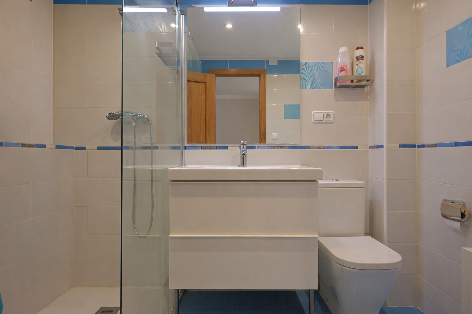 Badkamer met witte tegels, blauwe accenten, spiegel, moderne wastafel en glazen douchecabine.