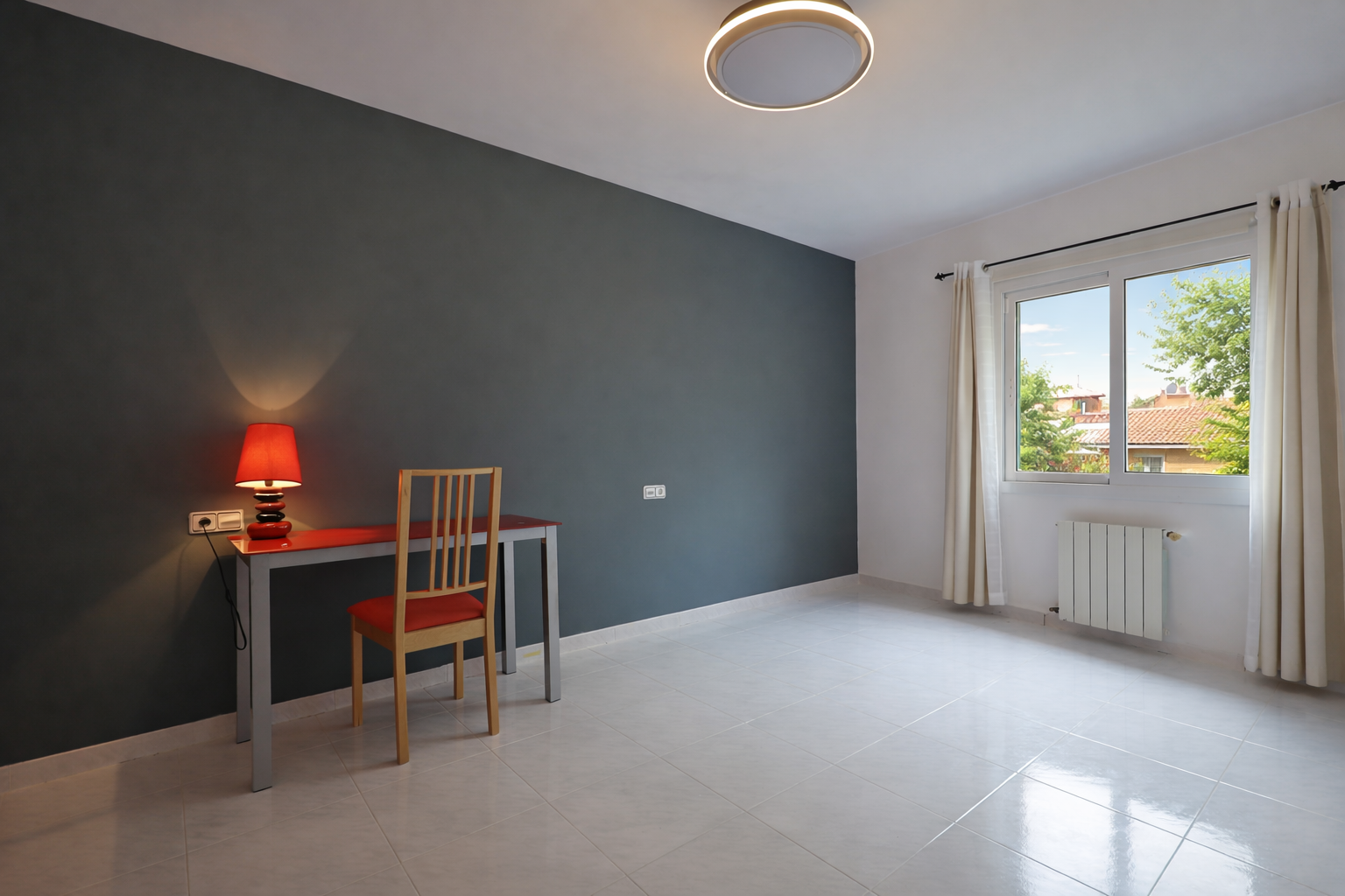Lichte kamer met rood bureau en loodgrijze muur, crèmekleurige vloertegels en een ronde plafondlamp.