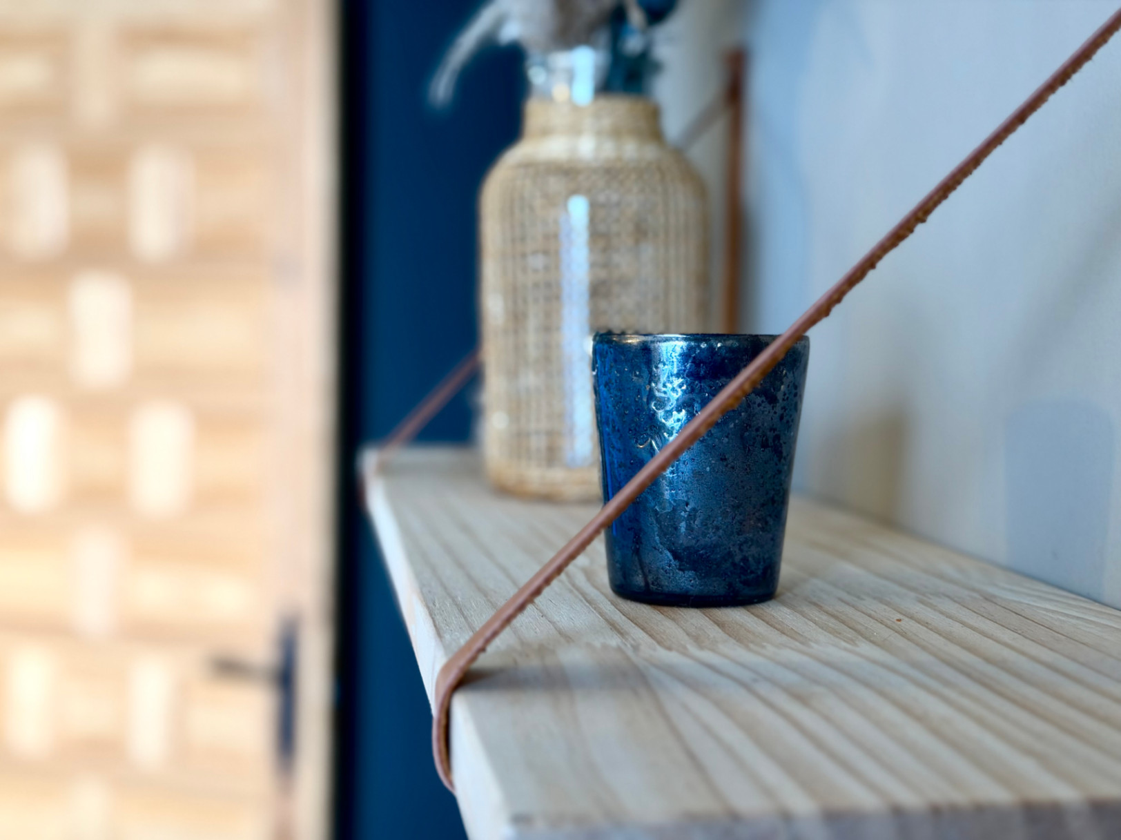 Close-up van een licht houten plank met decor: een getextureerd blauw glas en een glazen fles met een rieten hoes.