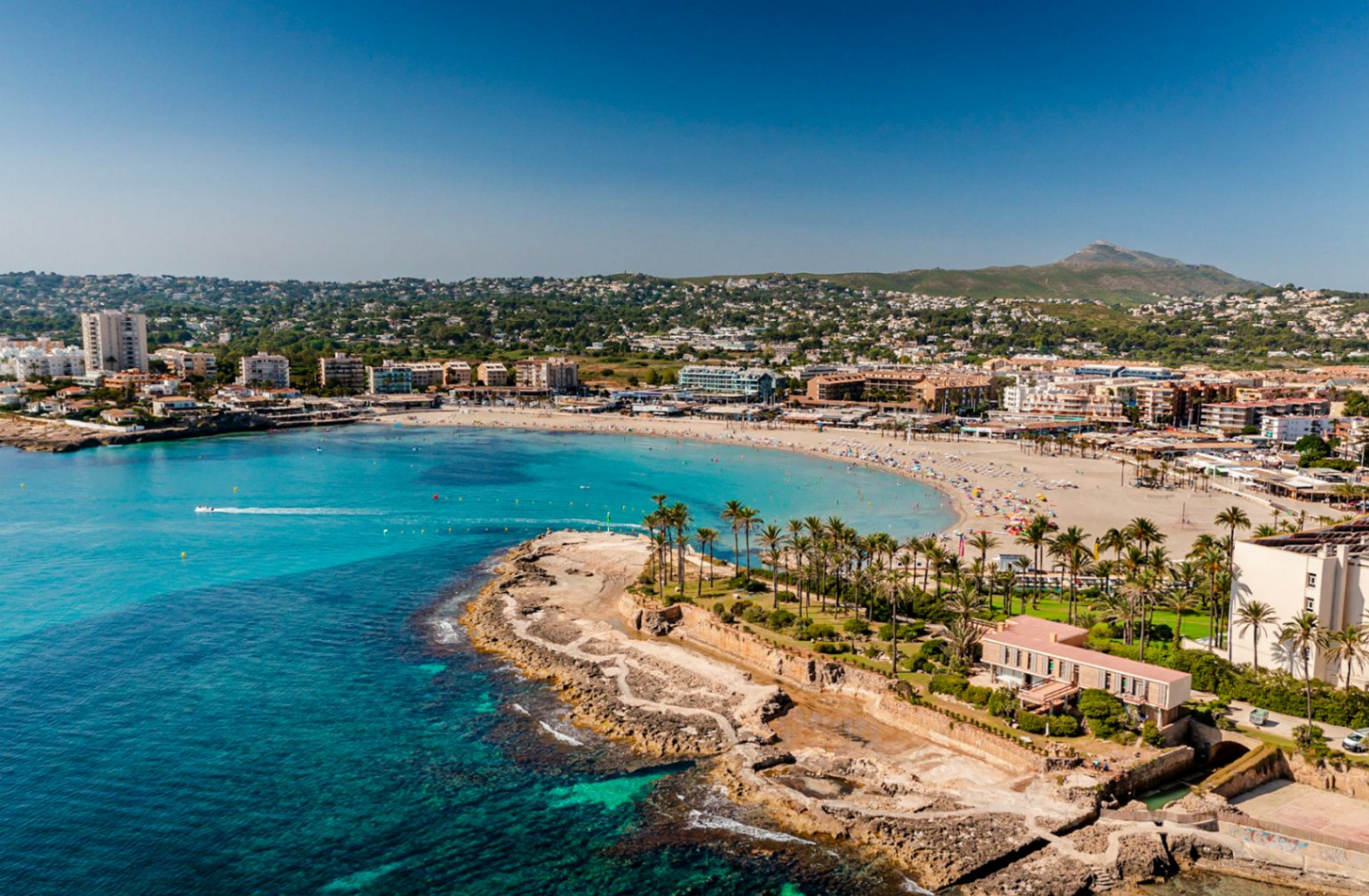 Luchtfoto van Playa El Arenal in Jávea, met zijn rotsachtige kustlijn en omliggende gebouwen onder een heldere hemel.