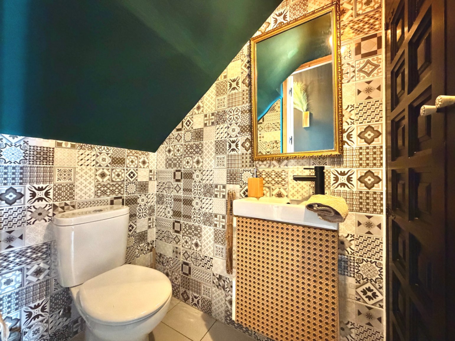 Toilet met patroontegels, een donkergroen plafond en een wastafelmeubel met rieten voorkant.